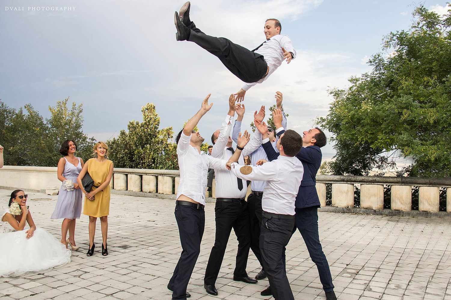 Wedding party members jump and pose playfully on a stone patio.