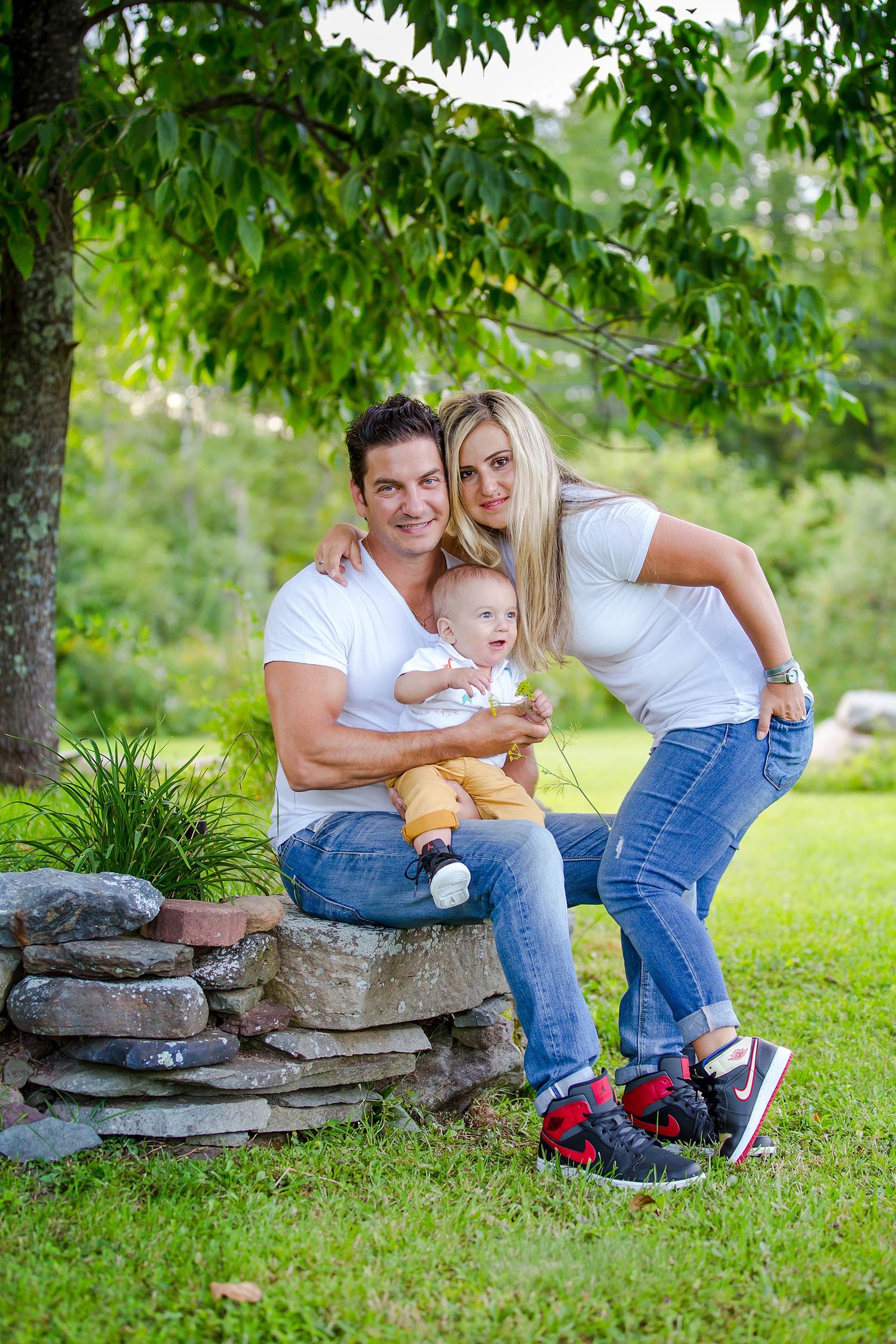 Family sitting on stone wall surrounded by green trees during outdoor photo session.