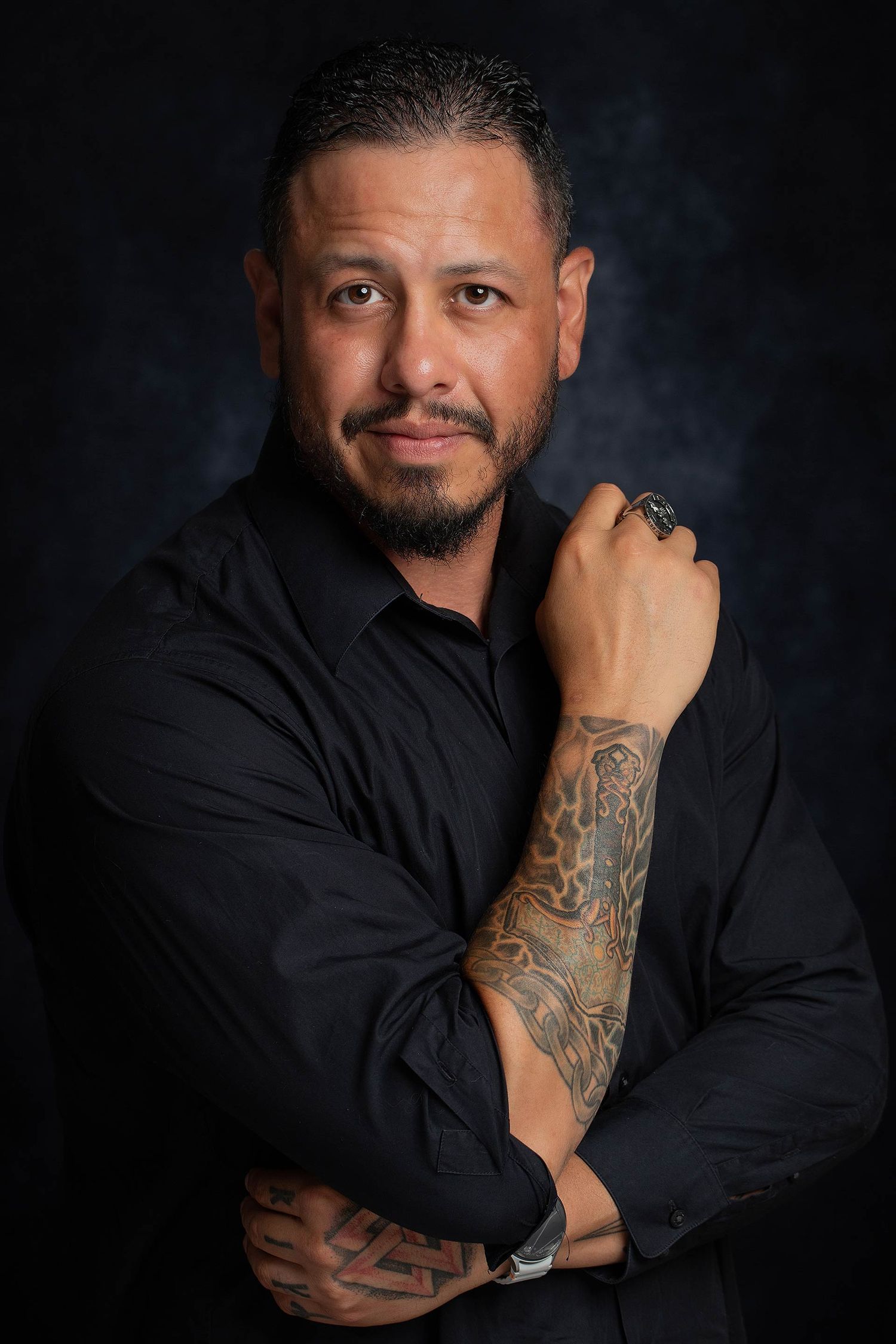 Professional portrait in black shirt showing tattooed arm against dark background with contemplative expression.
