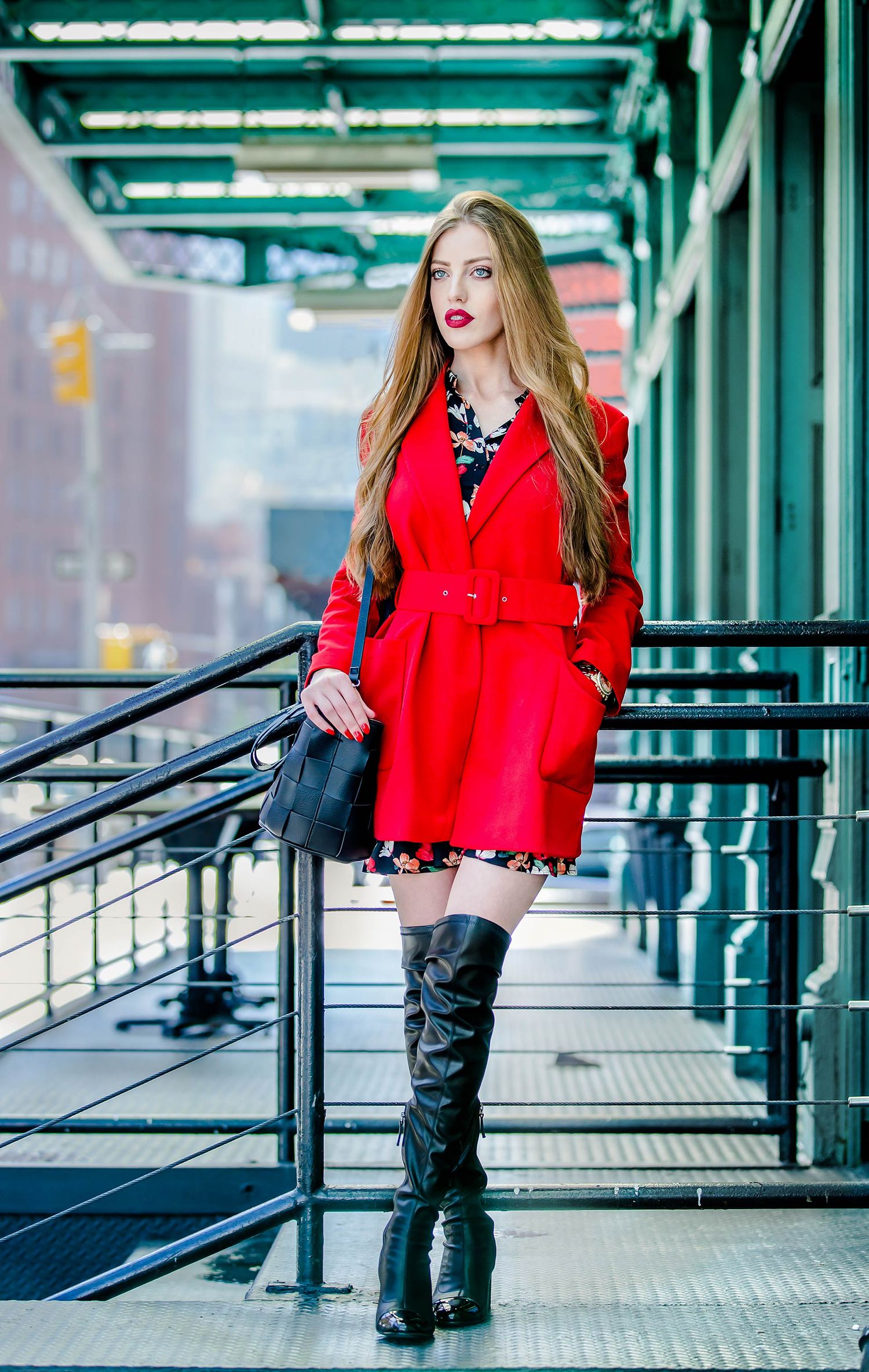 Fashion model in red coat and tall boots poses on city sidewalk with modern architecture.