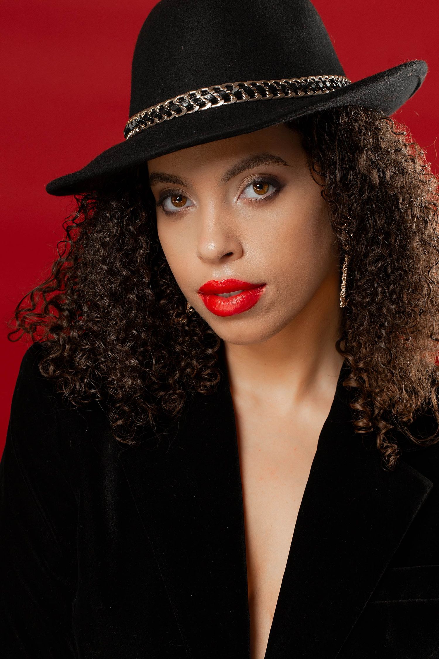Glamorous fashion portrait with black hat and velvet blazer against red background.