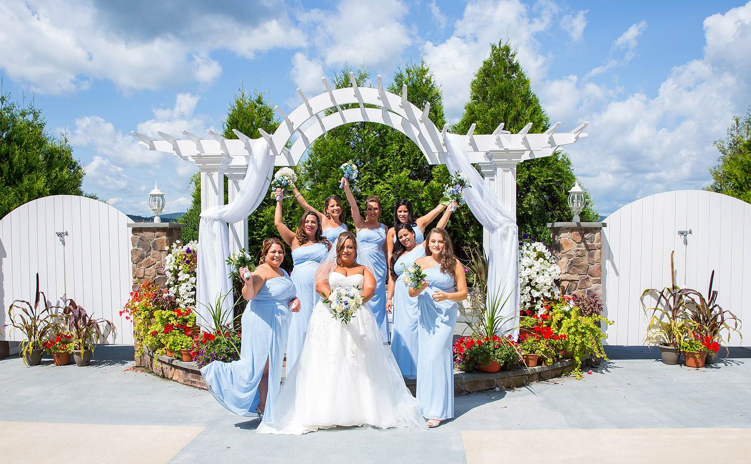 Bridesmaids in light blue dresses celebrating with bride under white garden arch.