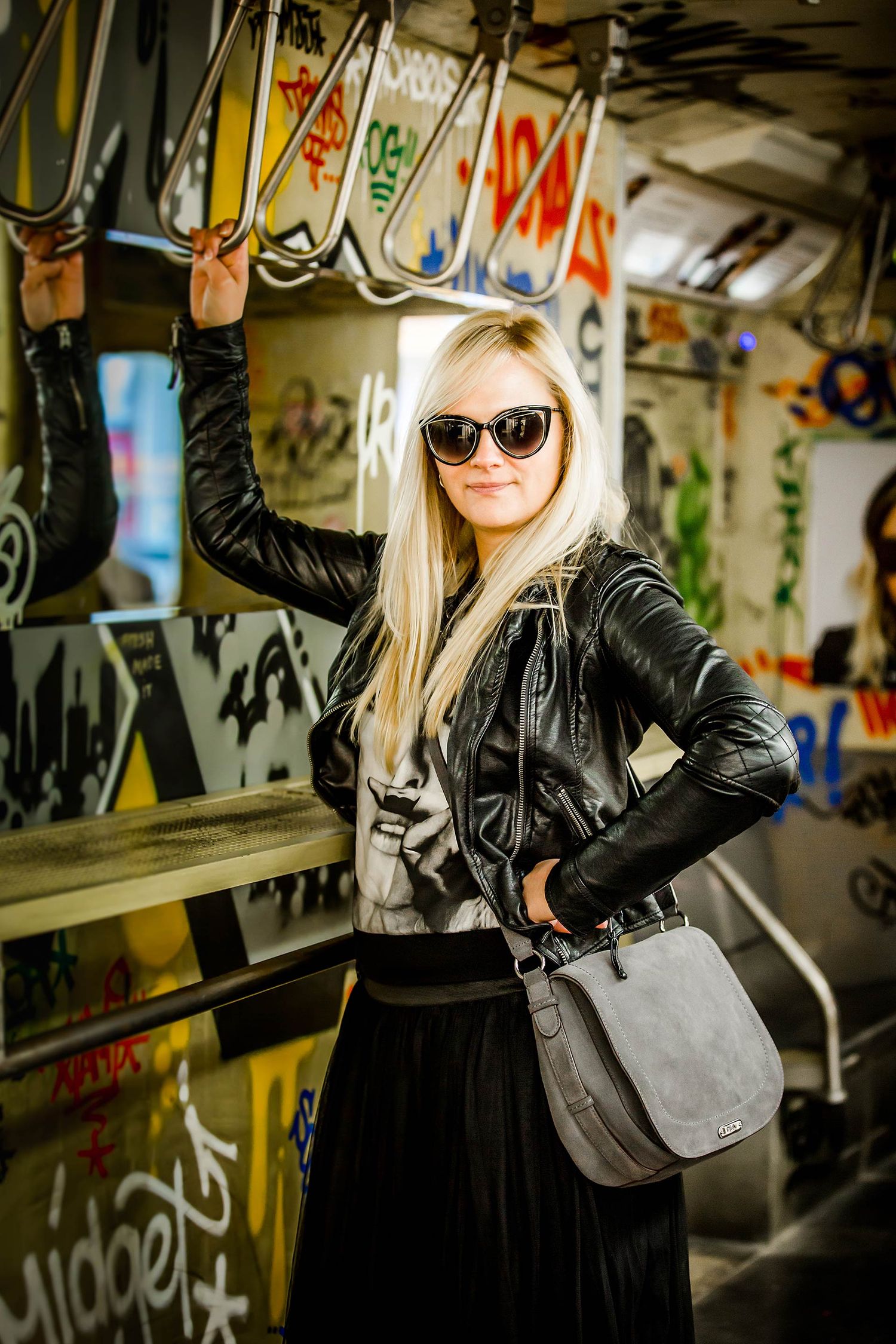 Stylish woman in leather jacket and sunglasses poses in urban graffiti setting.