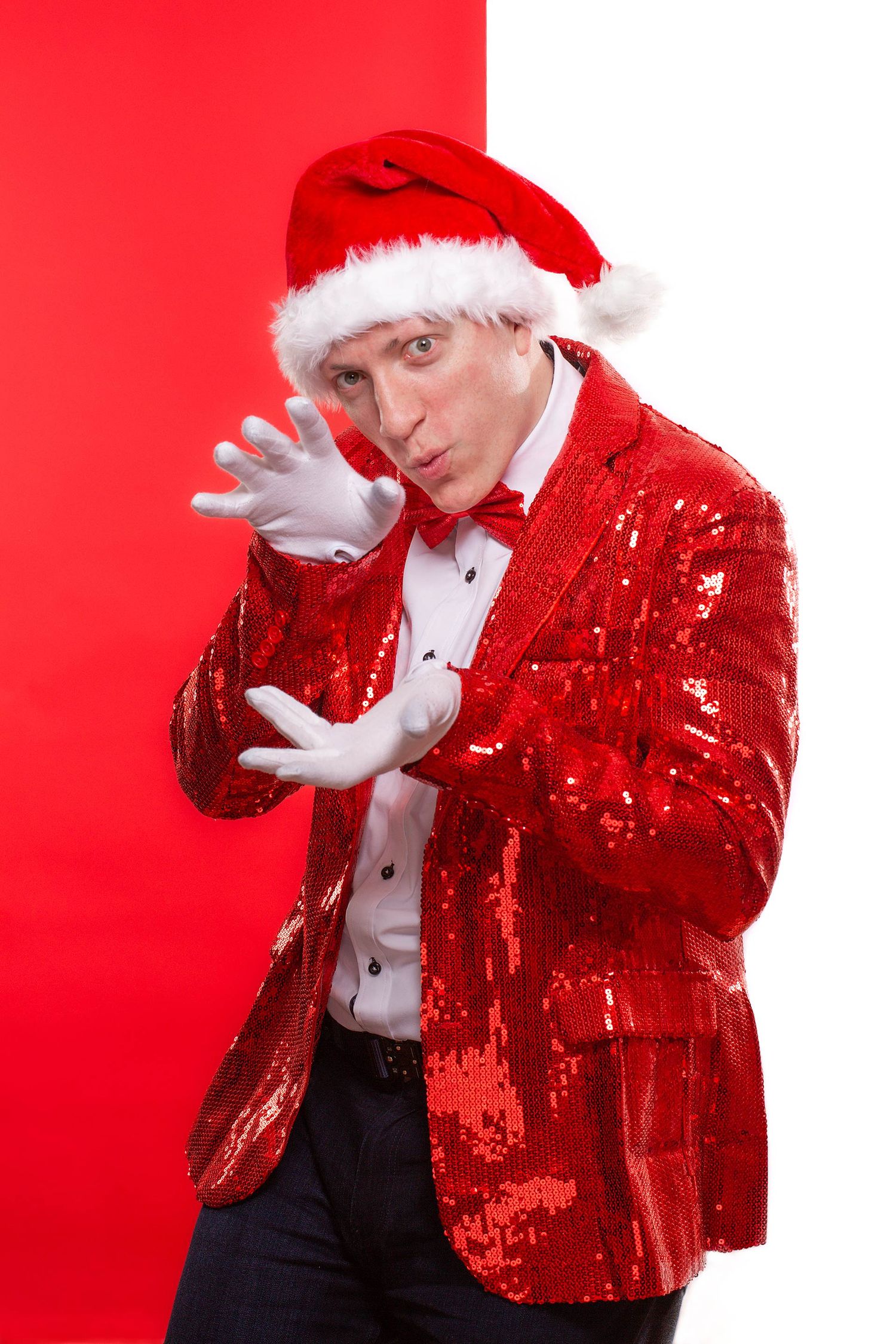Performer in red sequin jacket and Santa hat striking theatrical pose against red background.
