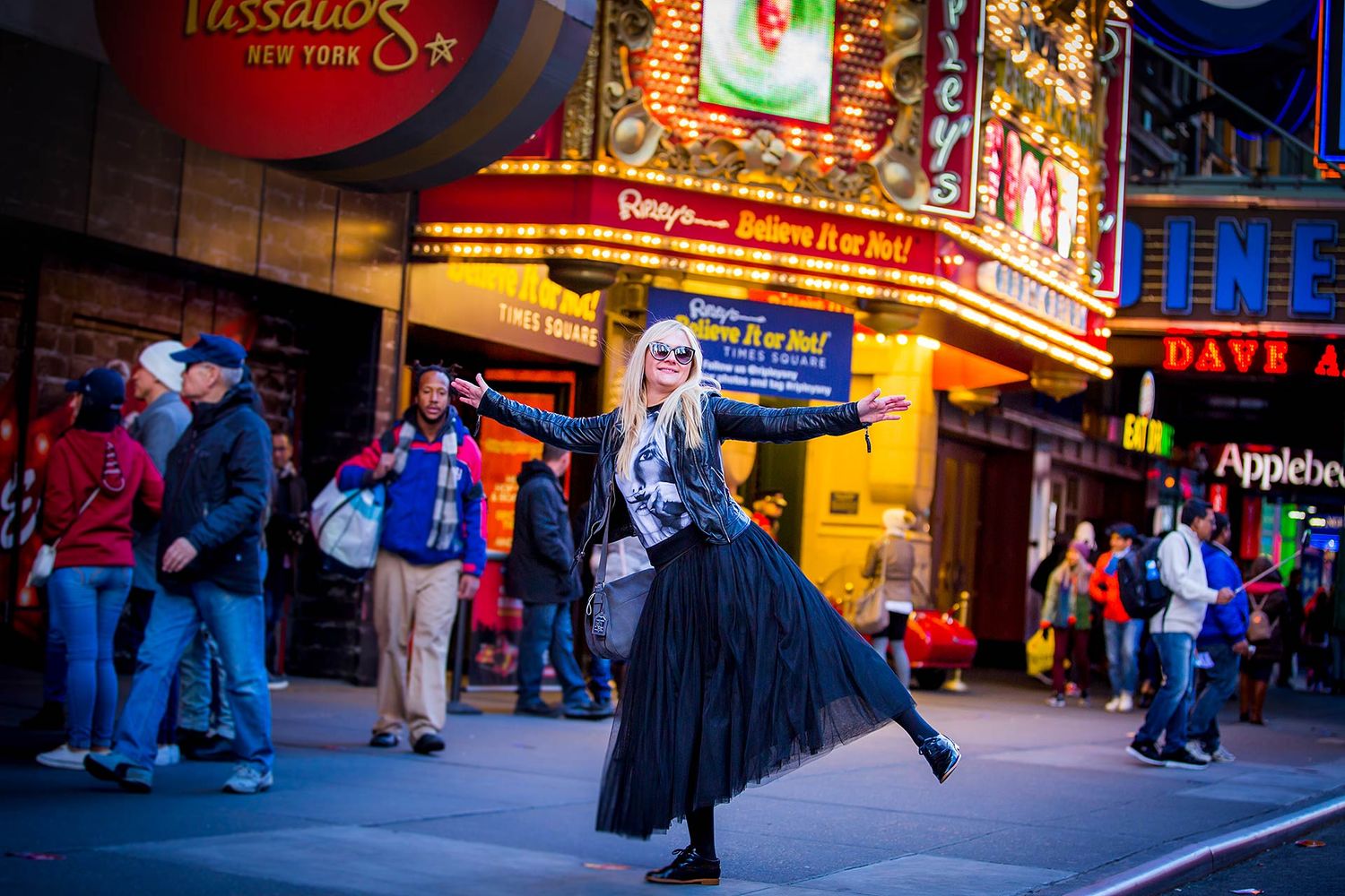 Woman in flowing black dress dances on city street at night near illuminated theater signs.