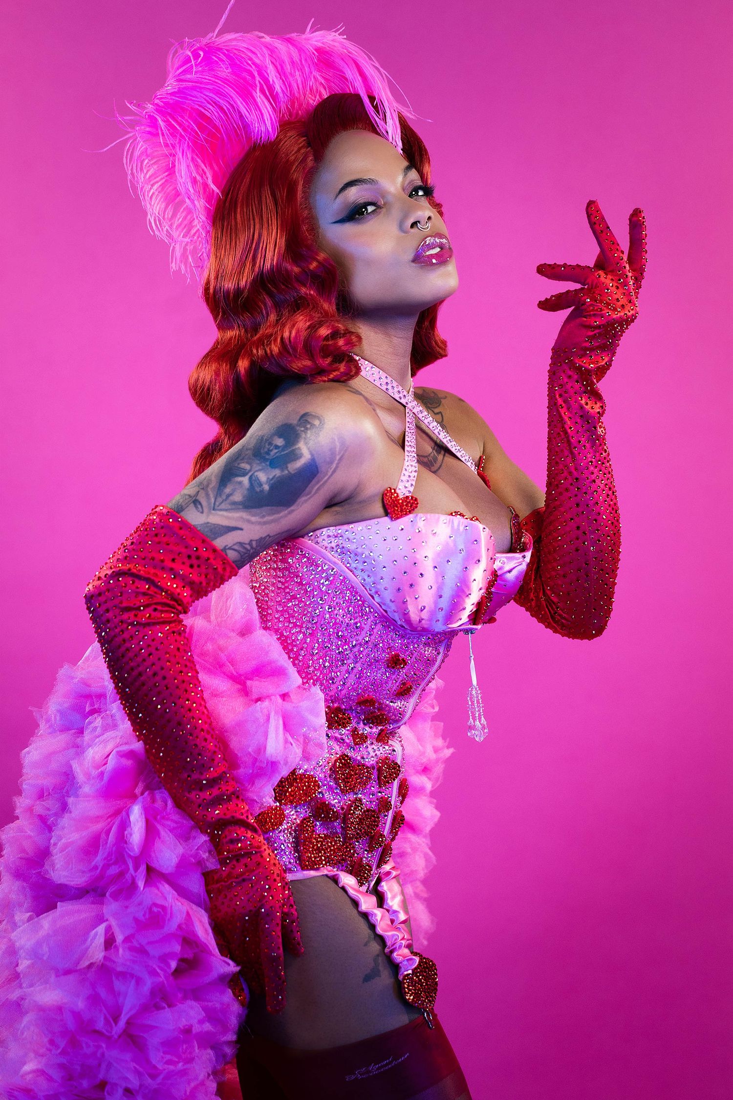 Burlesque performer in glittery costume poses against vibrant pink background.