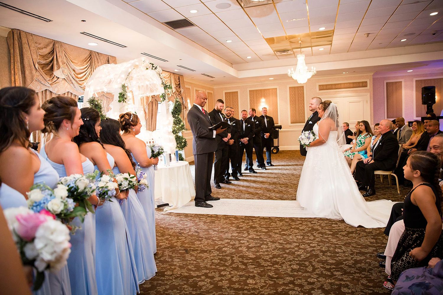 Indoor wedding ceremony with bridesmaids in blue and formal decor.