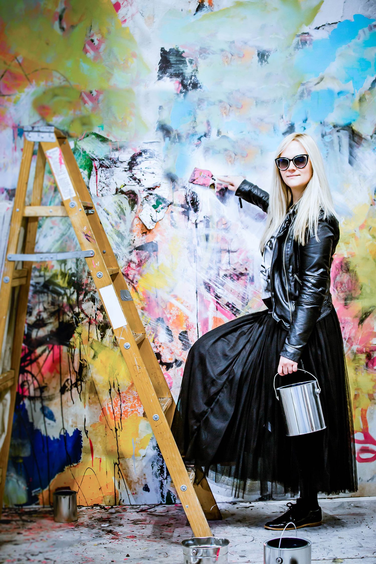 Fashionable woman in black leather jacket and tulle skirt poses with paint bucket against colorful graffiti wall.