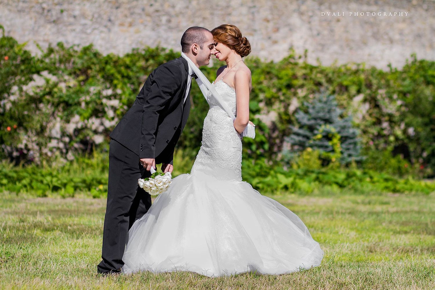 A wedding couple poses romantically in an outdoor garden setting with stone walls and greenery.
