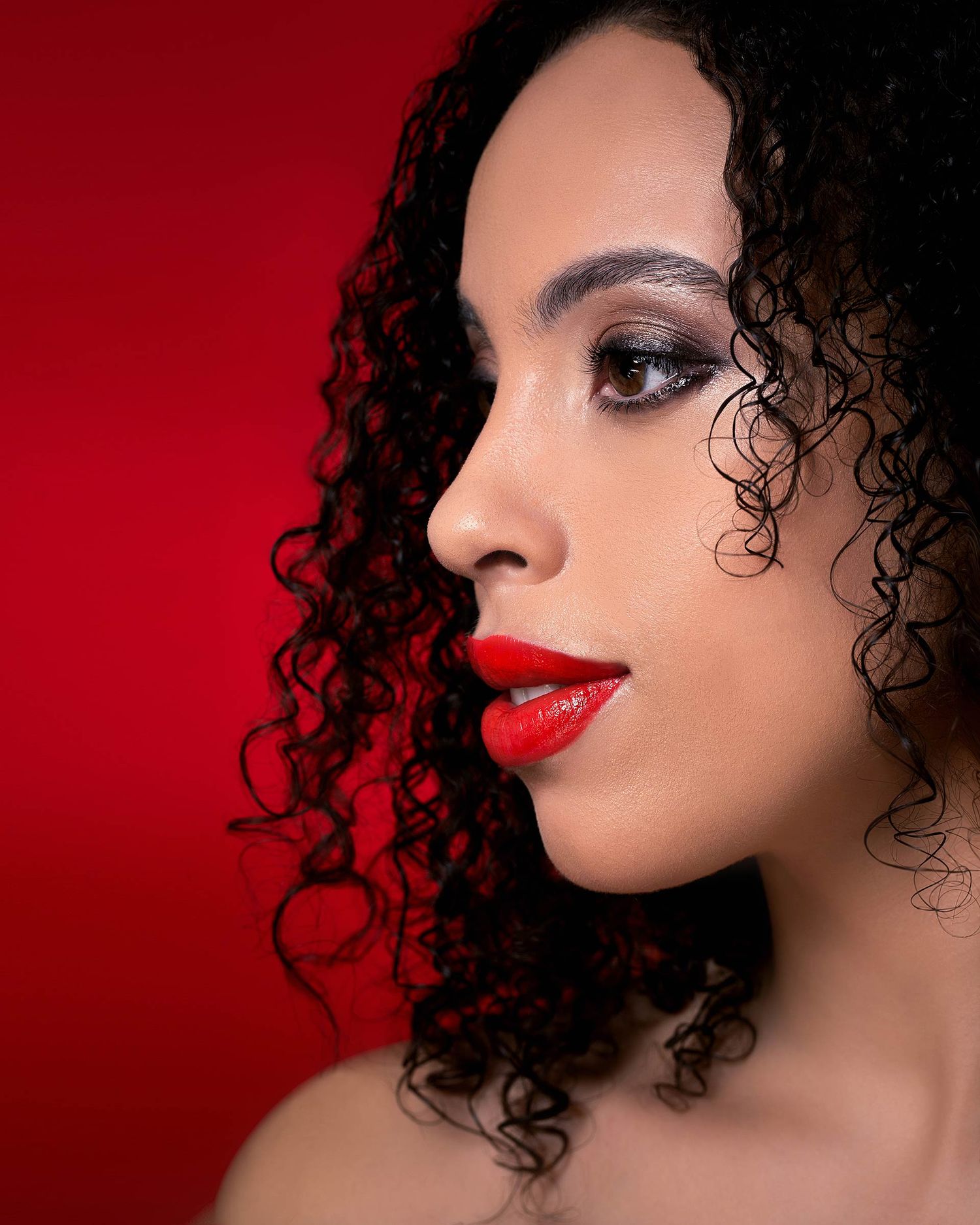 Glamorous beauty portrait with curly hair, smokey eyes and red lips against red background.