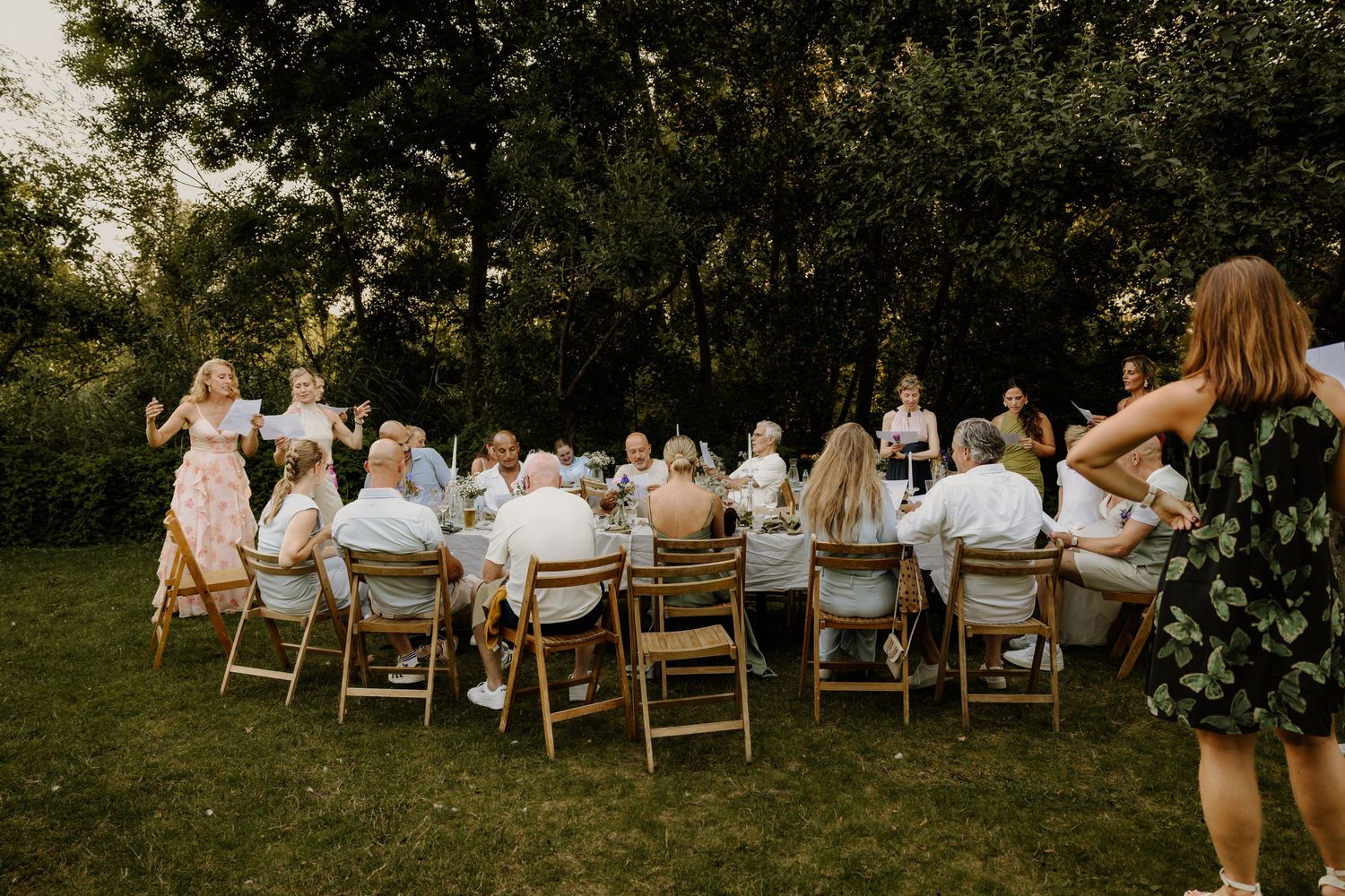 Gasten zitten aan lange tafels tijdens een diner in de buitenlucht en zingen samen met de zussen van de bruid een lied voor het bruidspaar.