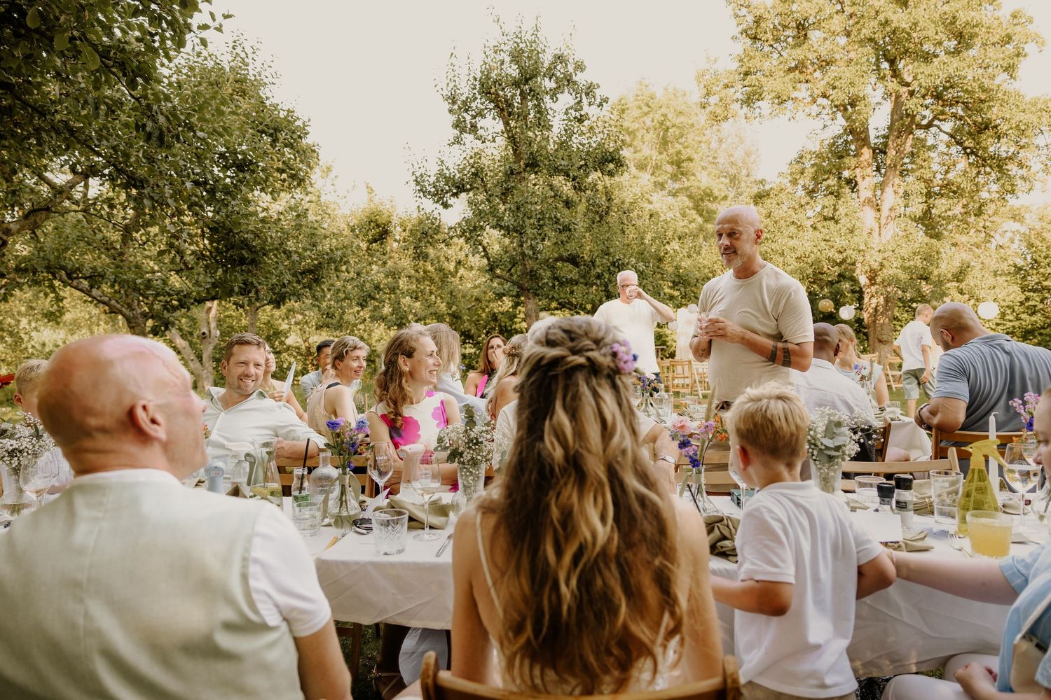 Gezellig bruiloftsdiner aan lange tafels tussen het groen, terwijl papa een speech houd.