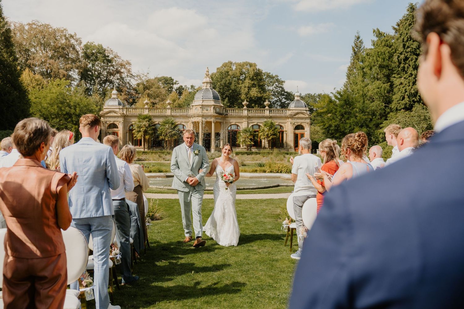 Bruid wordt door haar vader naar de ceremonie gebracht terwijl de gasten toekijken bij Oranjerie Hydepark