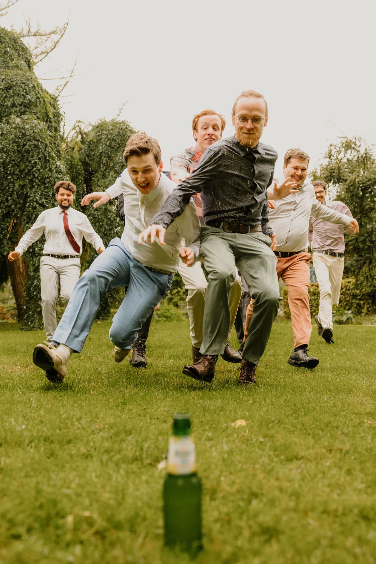 De bruidegom rent lachend samen met vrienden over het gras richting een flesje bier