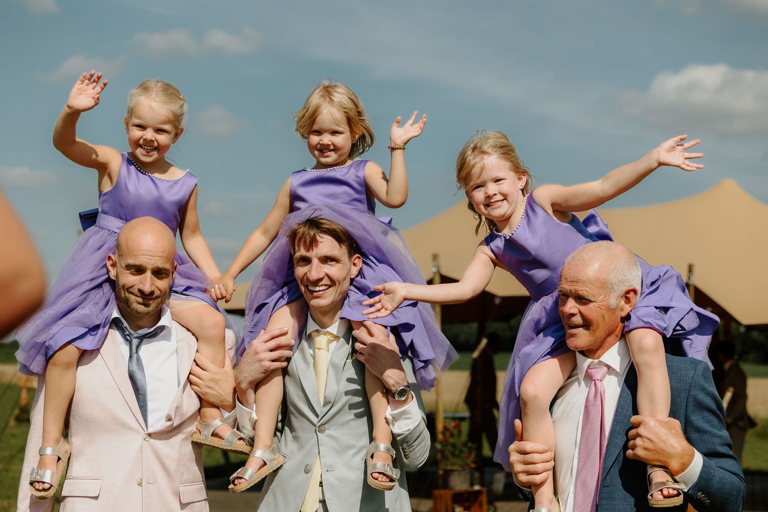 Drie jonge bruidsmeisjes in paarse jurkjes zwaaien vanaf de schouders van hun vader, oom en opa op een zomerse buitenbruiloft.