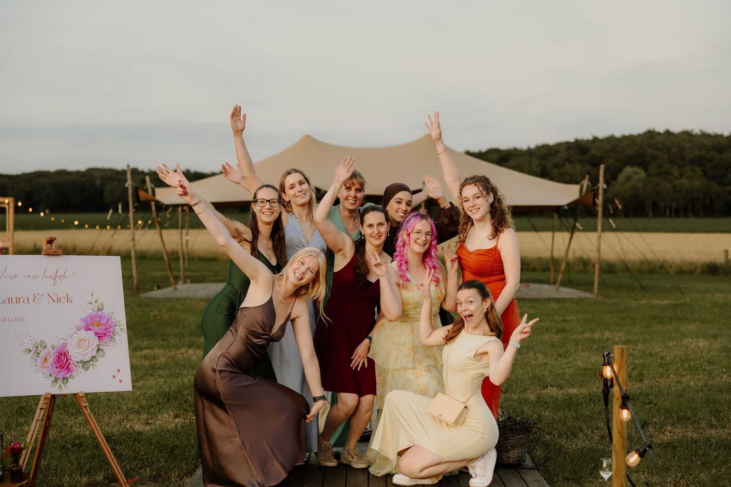 Bruiloftsgasten poseren blij voor een groepsfoto bij het welkomstbord op een zomerse buitenbruiloft