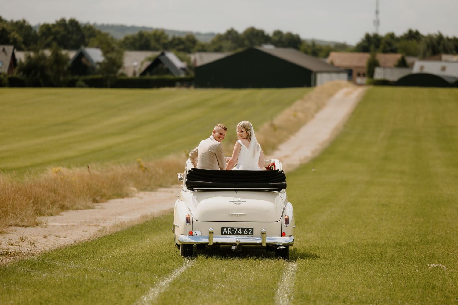 een bruidspaar zit vrolijk lachend in de trouwauto terwijl ze over een weiland rijden.