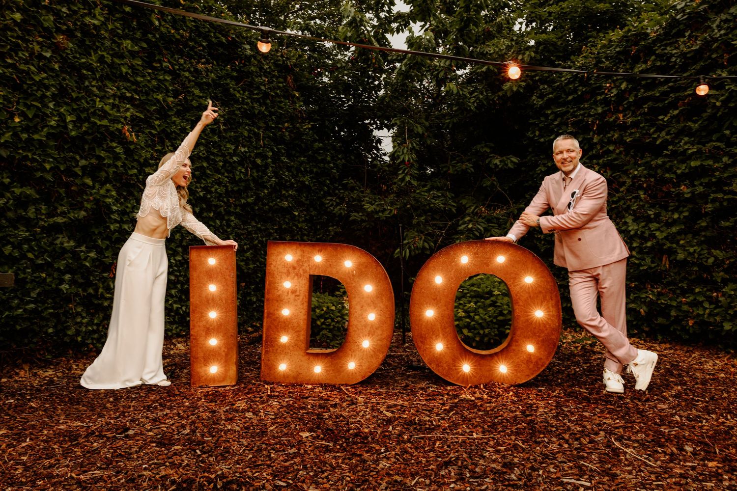Bruidspaar bij verlichte I DO letters op hun trouwlocatie