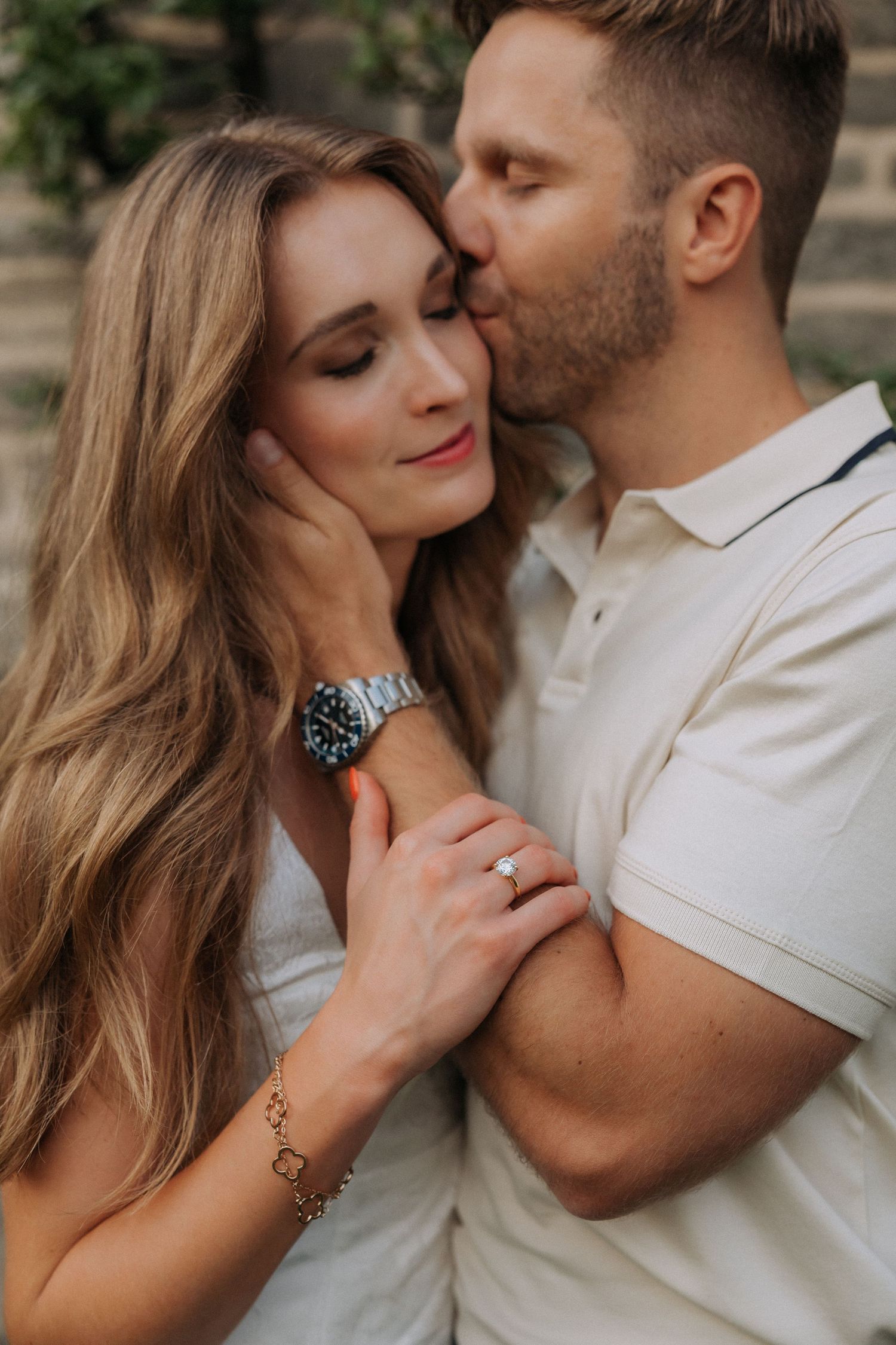 Man tenderly kissing woman on cheek while embracing her, she displays engagement ring and watch.