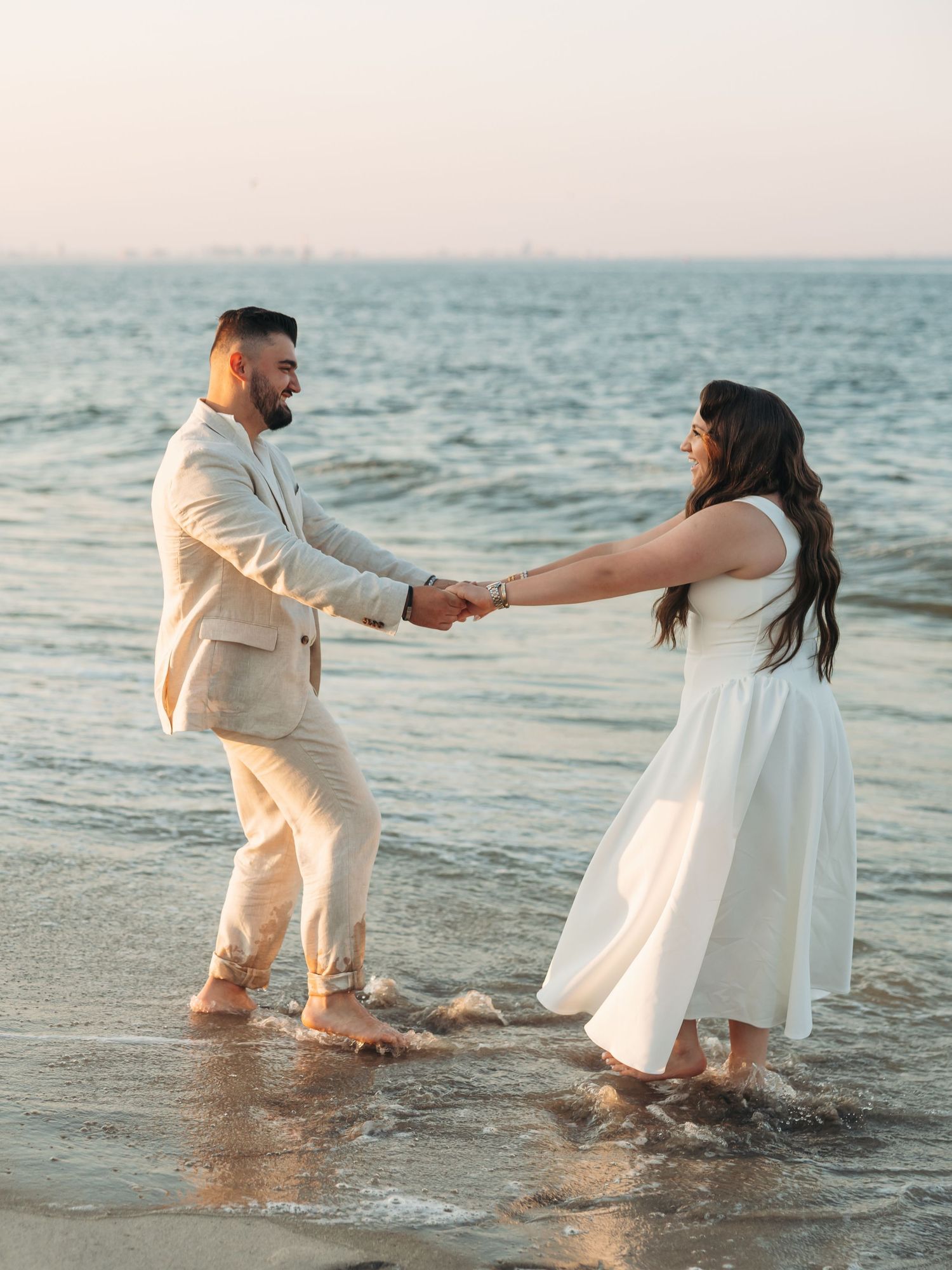 Couple holding hands and dancing in ocean waves at sunset, wearing white dress and beige suit on sandy beach.