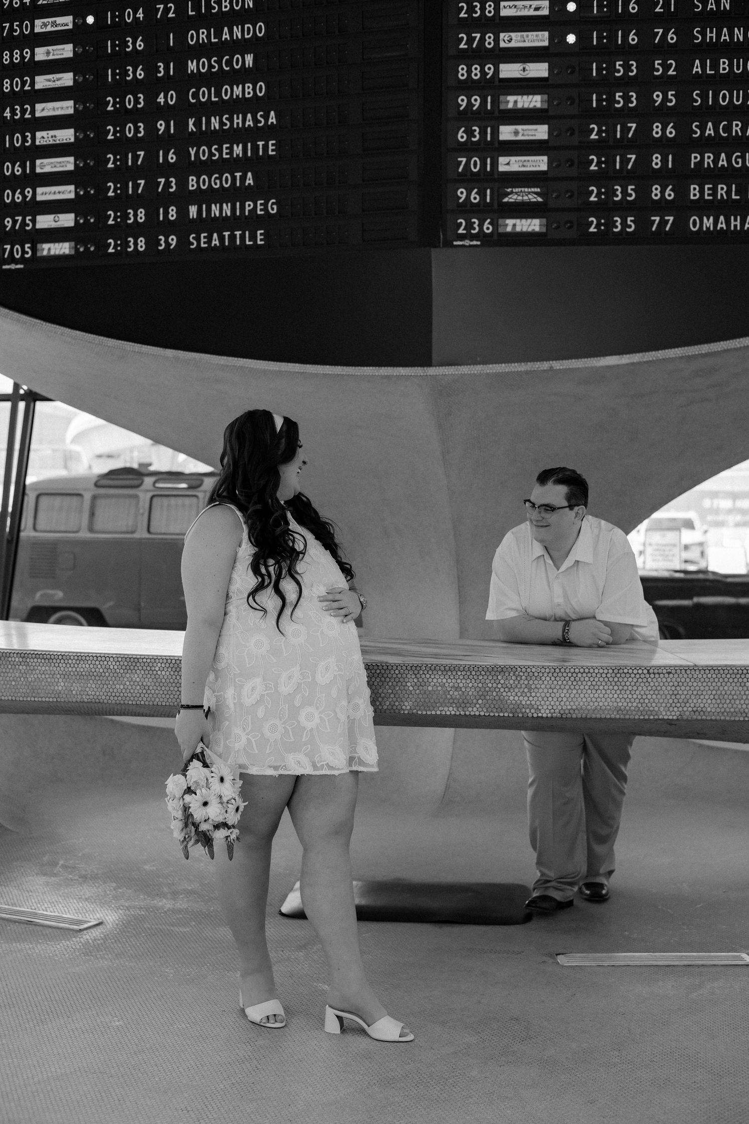 Black and white photo of pregnant bride in white dress holding bouquet at airport departure board.