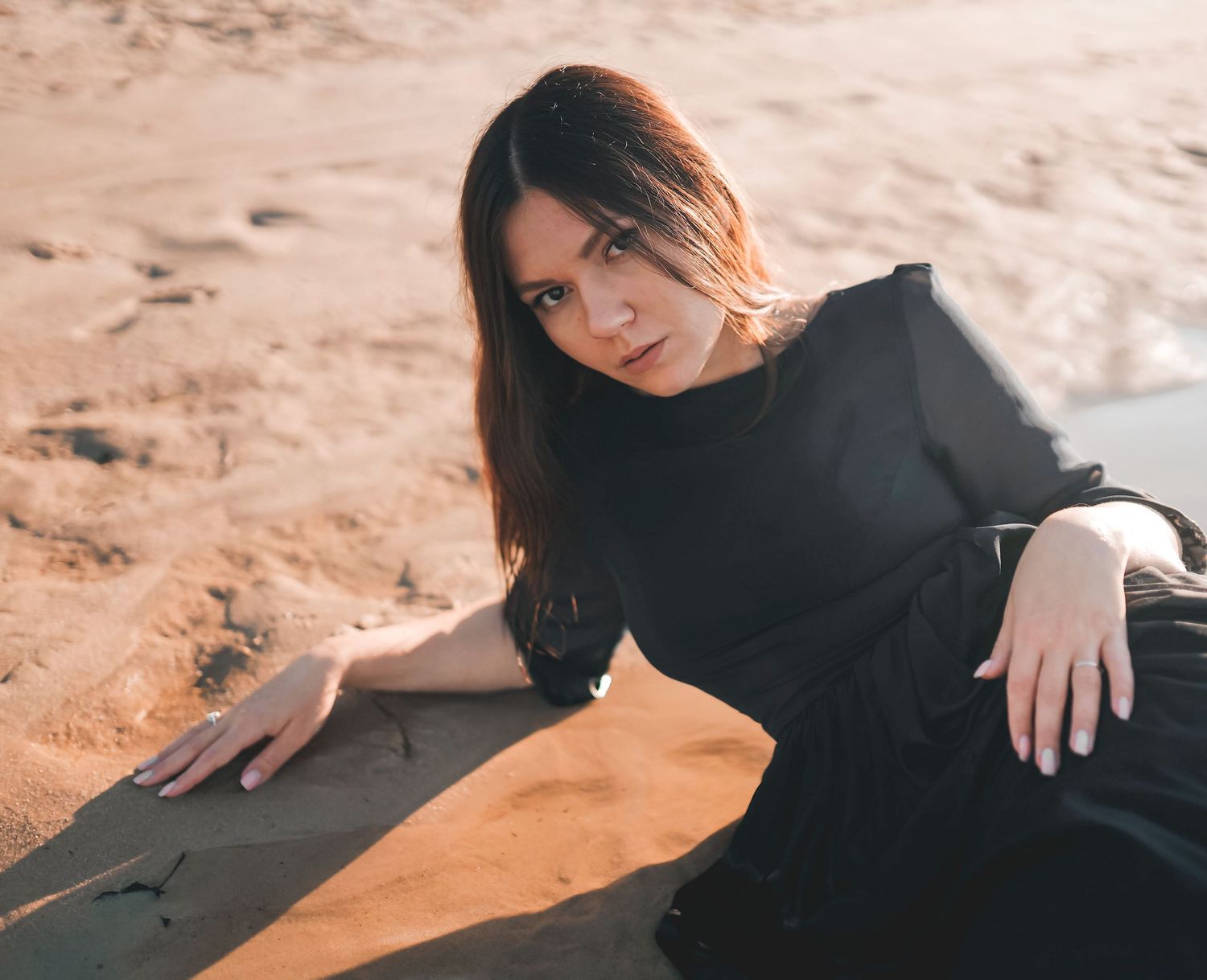 Young woman in black dress posing gracefully on sandy beach surface in warm golden sunlight.