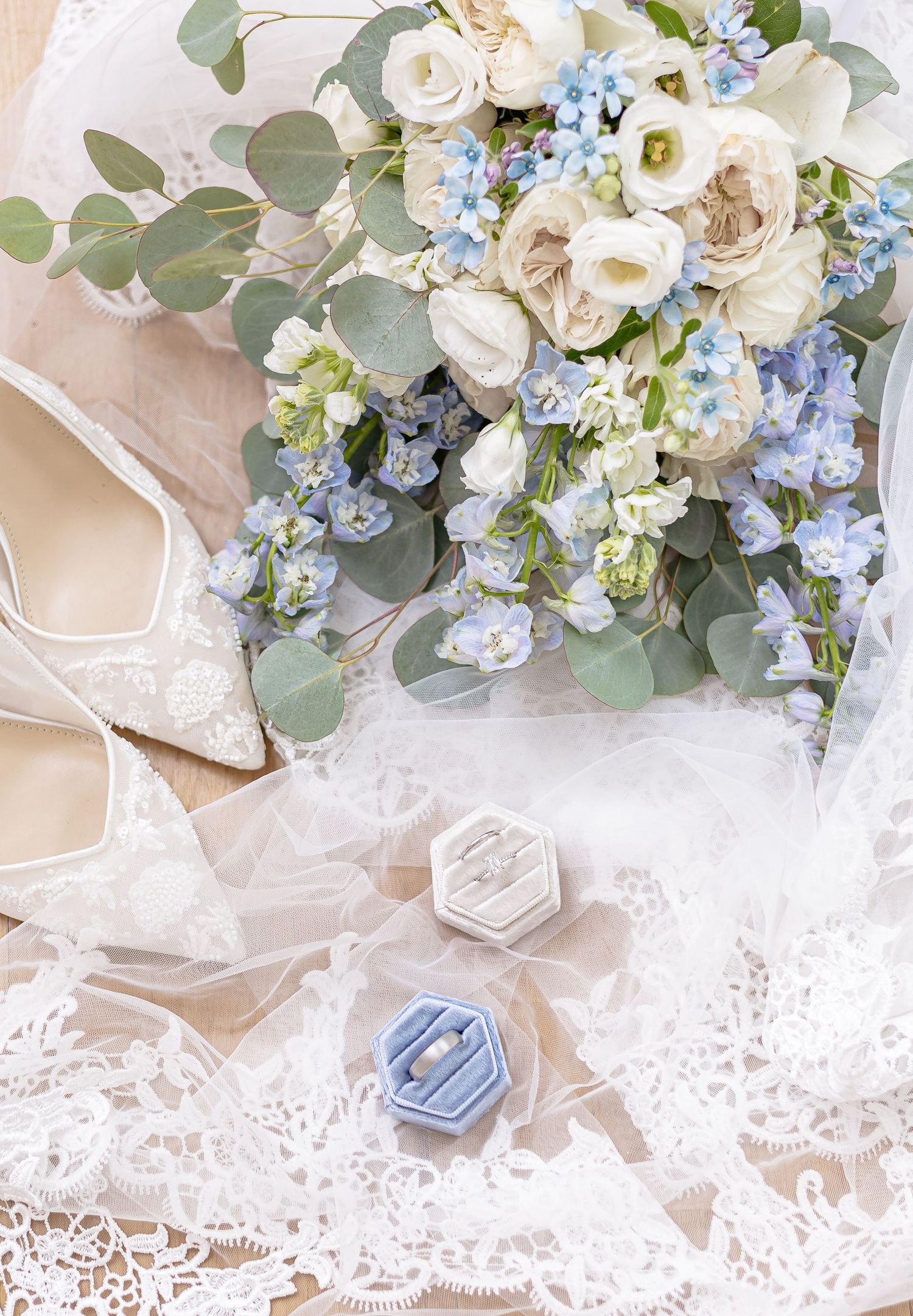 Wedding flat lay with white and blue bouquet, lace veil, heels, and ring boxes.