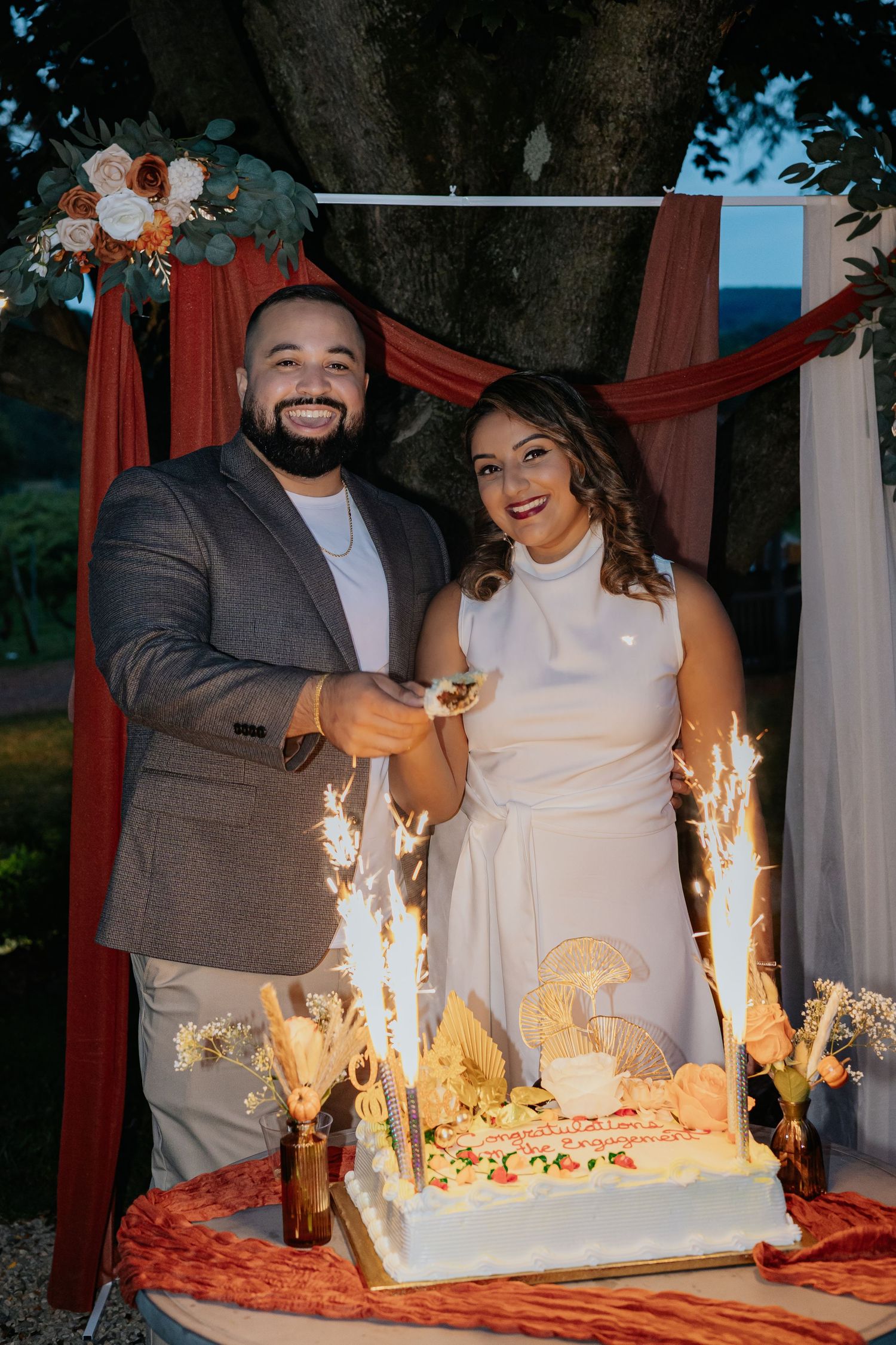 Smiling couple posing with sparkler-lit engagement cake under decorated outdoor arch.