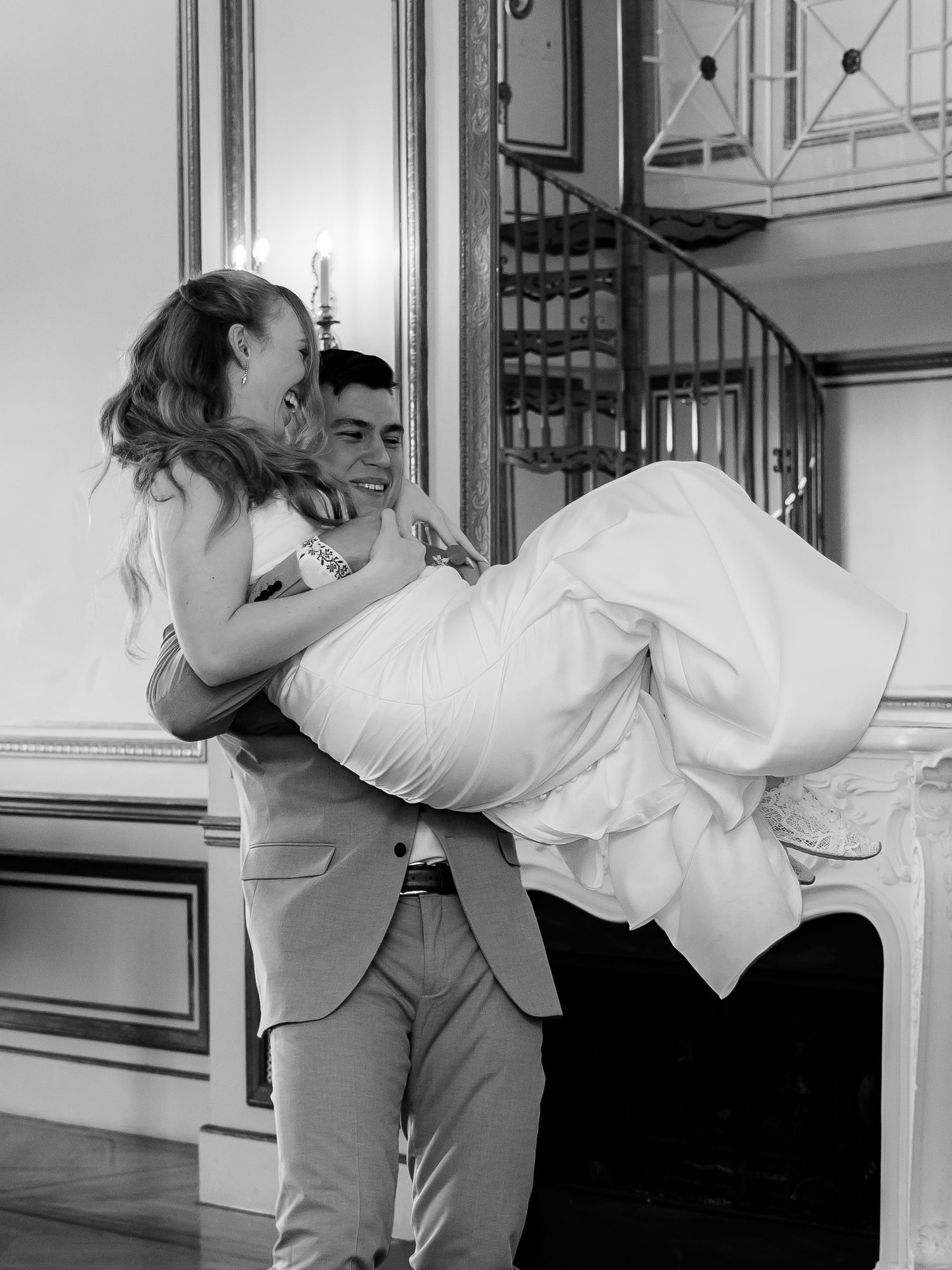 Black and white photo of groom carrying laughing bride in his arms in a grand ballroom.