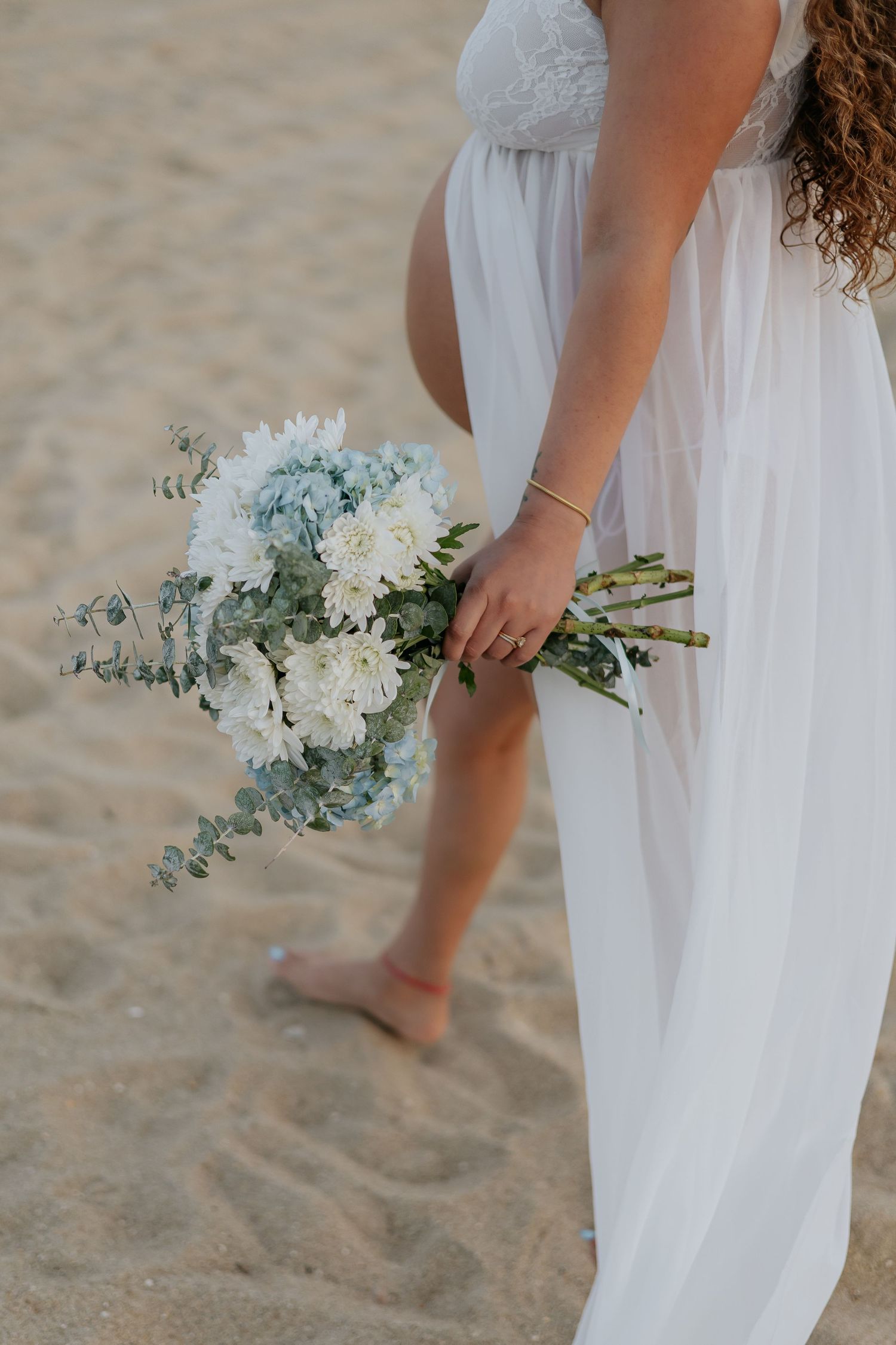 Pregnant woman in white dress holding blue and white flower bouquet during beach maternity session.