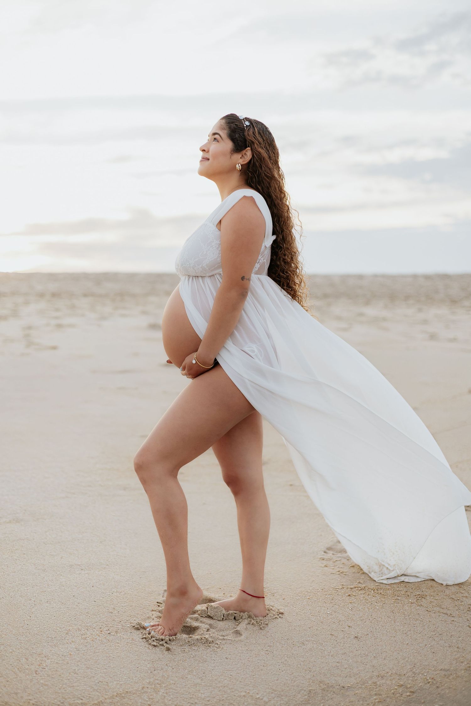 Pregnant woman in flowing white dress standing on open beach holding belly, looking upward at sky.