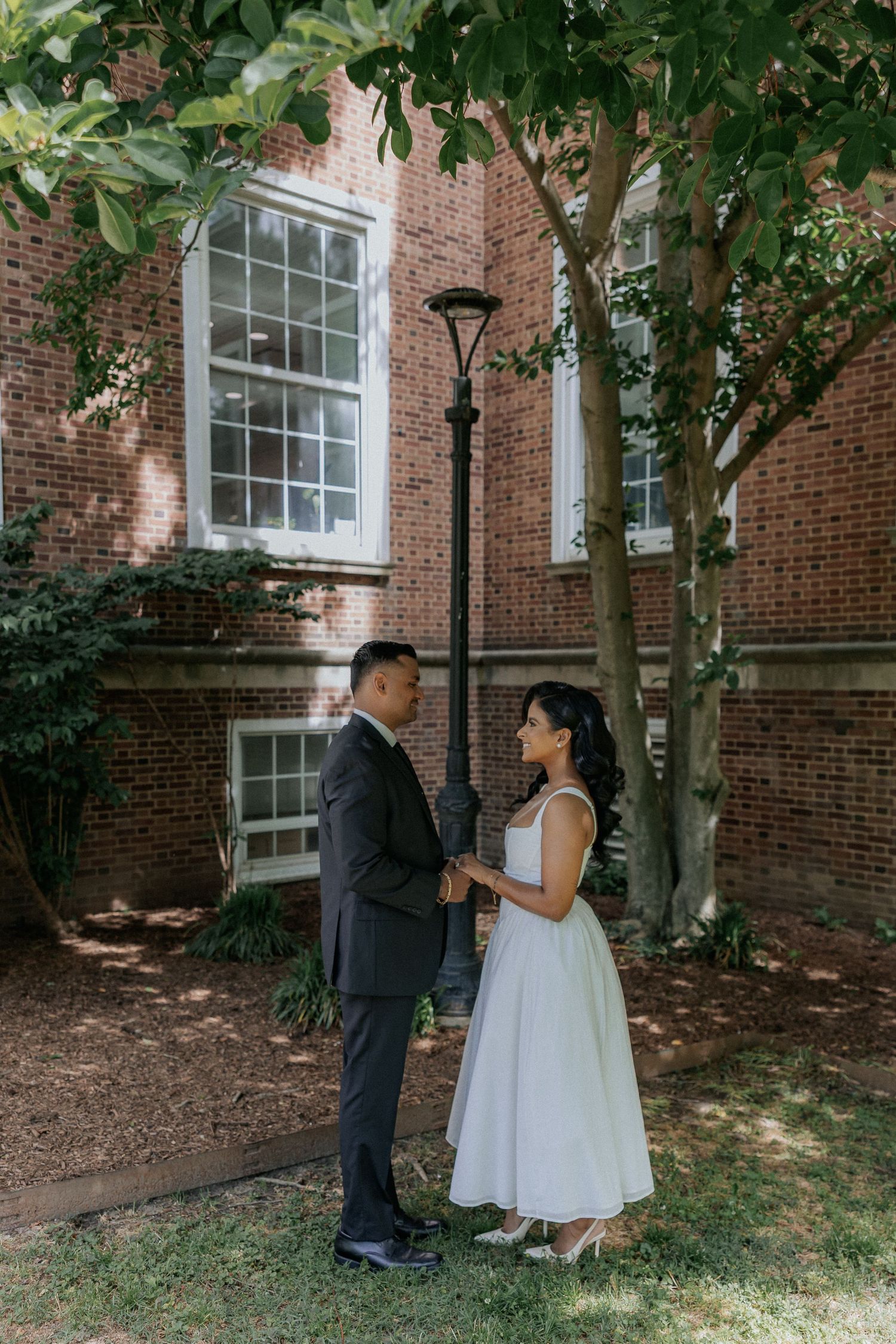 Bride and groom holding hands facing each other near brick building and lamppost in garden.