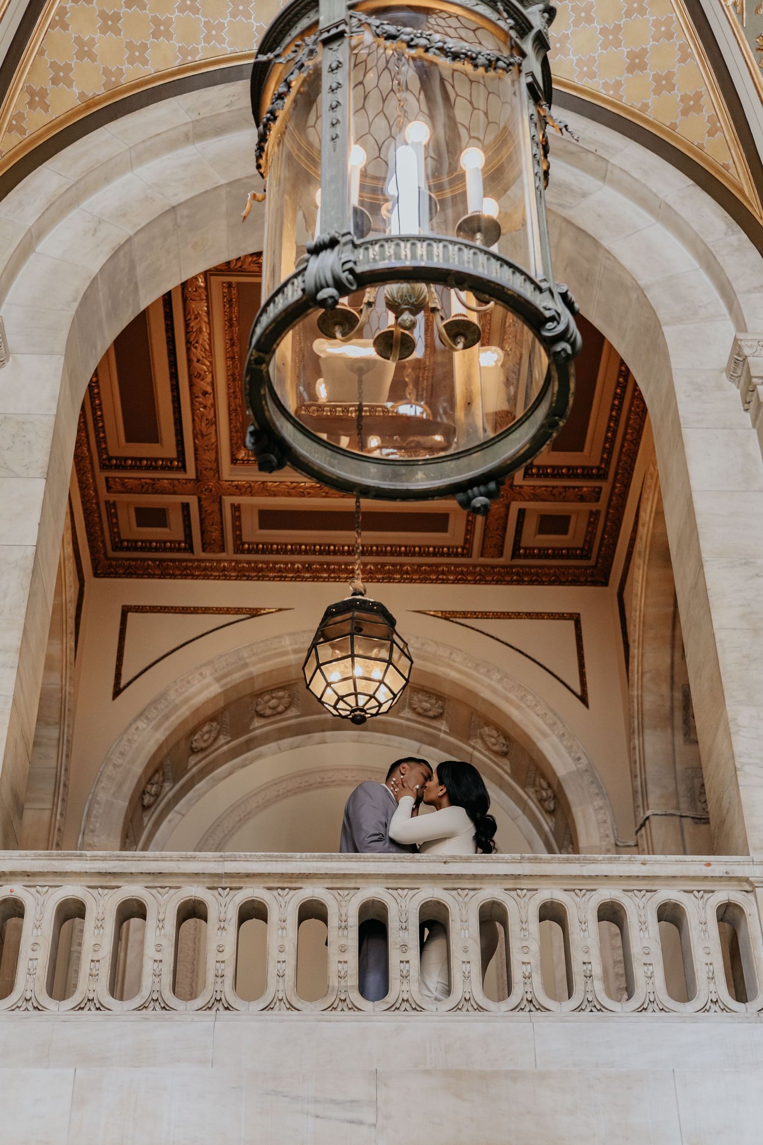 Couple kisses on balcony beneath ornate chandelier and gilded arched ceiling.