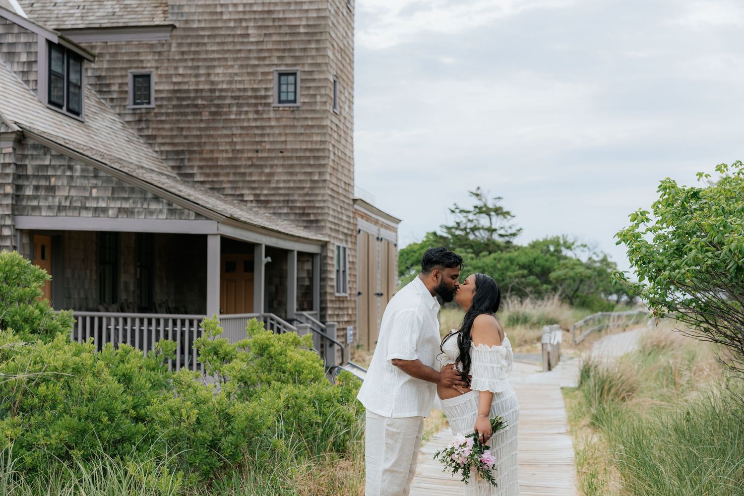 Couple shares romantic kiss on wooden boardwalk beside charming shingle cottage near sandy dunes.