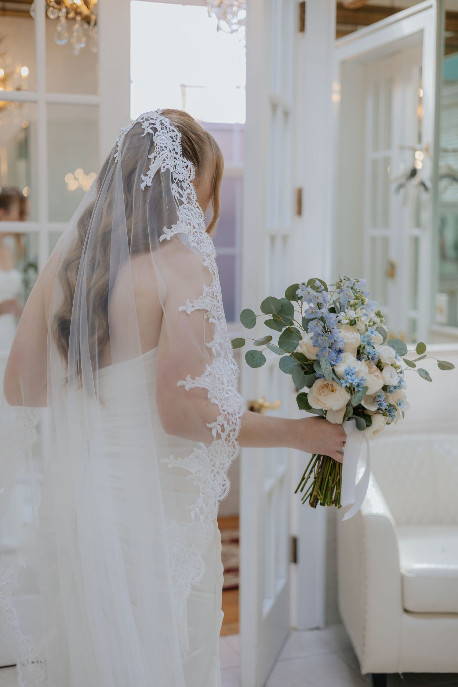 Bride in lace-trimmed veil holding blue and white eucalyptus bouquet standing by white French doors.
