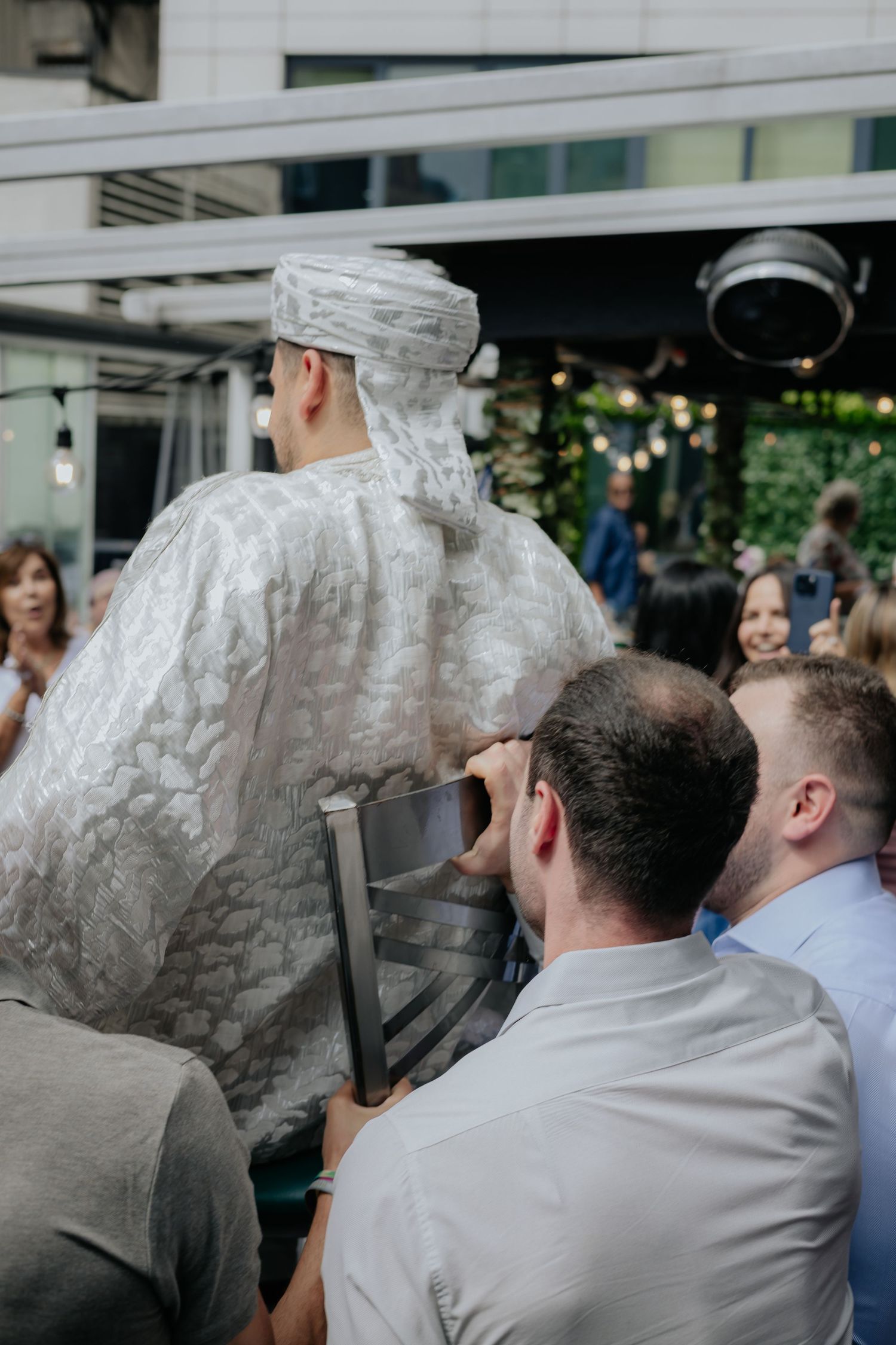 Groom in white traditional robe and turban is lifted on a chair by guests at a wedding celebration.