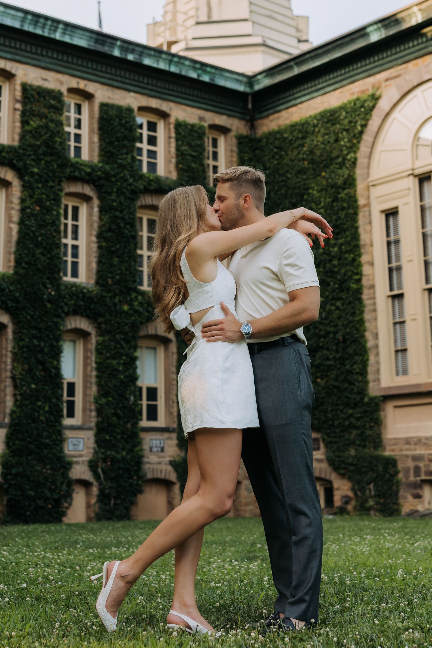 Couple sharing a romantic kiss on green lawn before ivy-covered historic brick building.