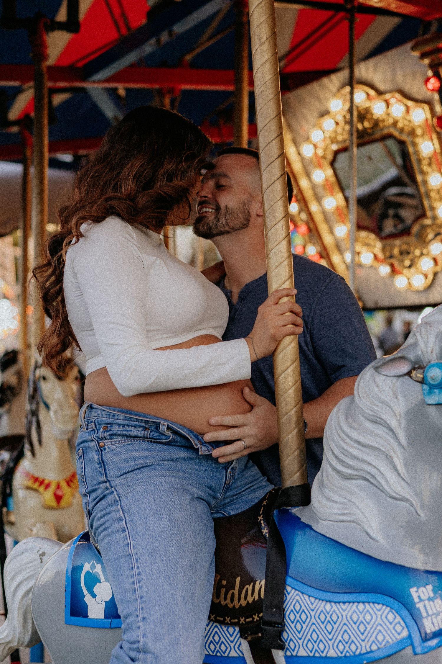 Expecting couple sharing a kiss on a carousel ride at a brightly lit amusement park.