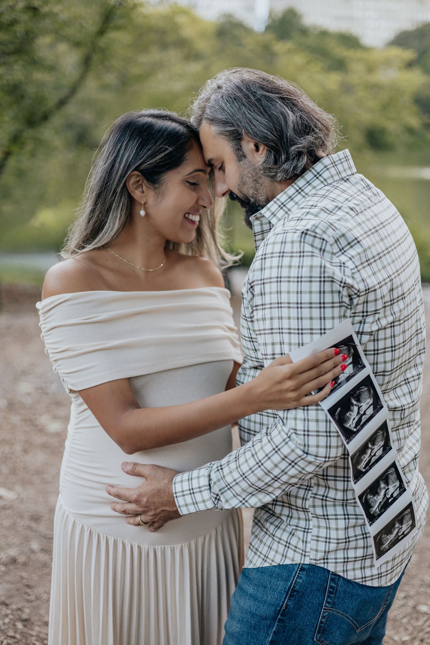 Expecting couple touching foreheads while man holds ultrasound photos in Central Park.