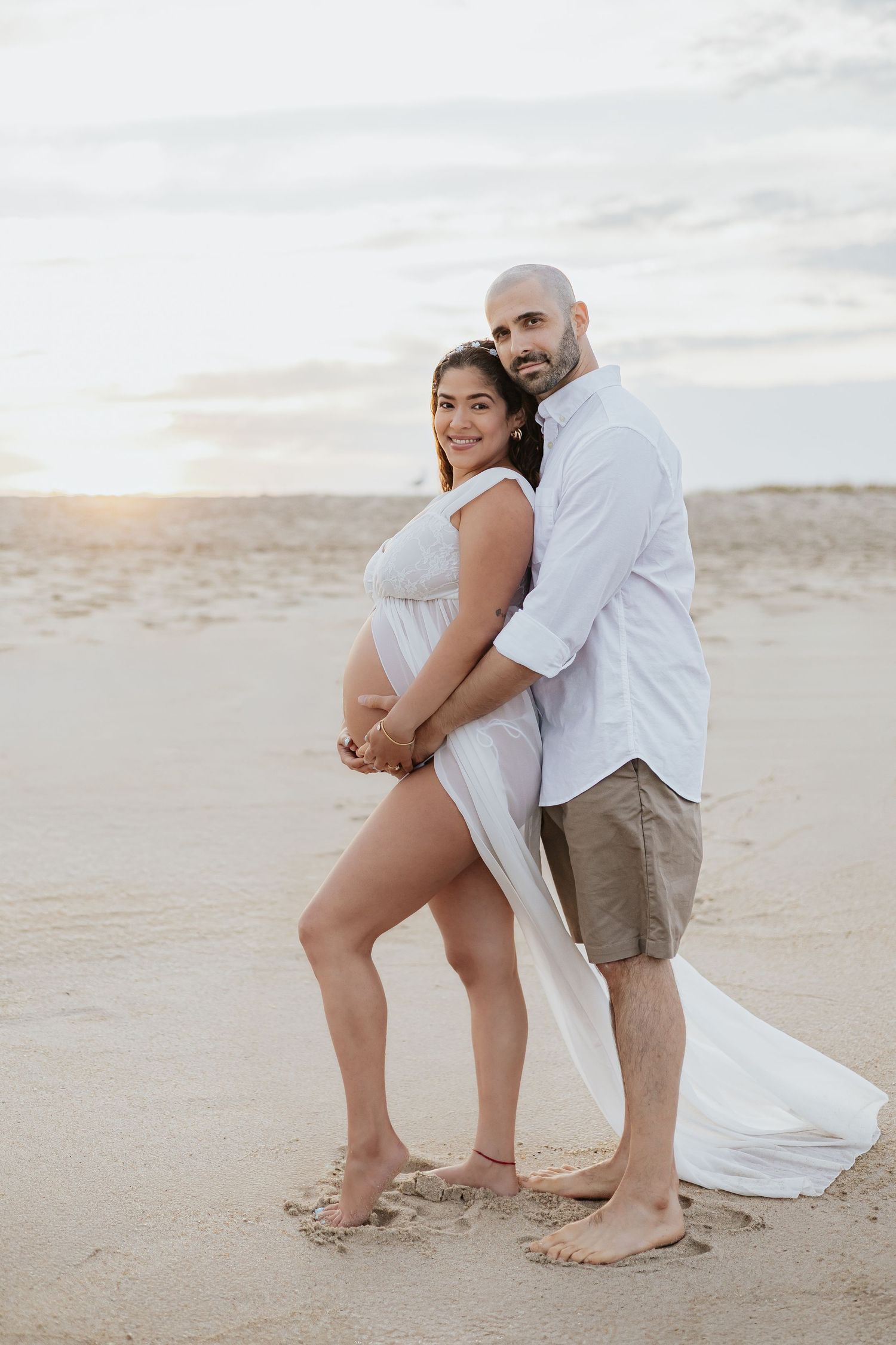 Pregnant couple posing at sunset on beach, man embracing woman in white flowing maternity dress.