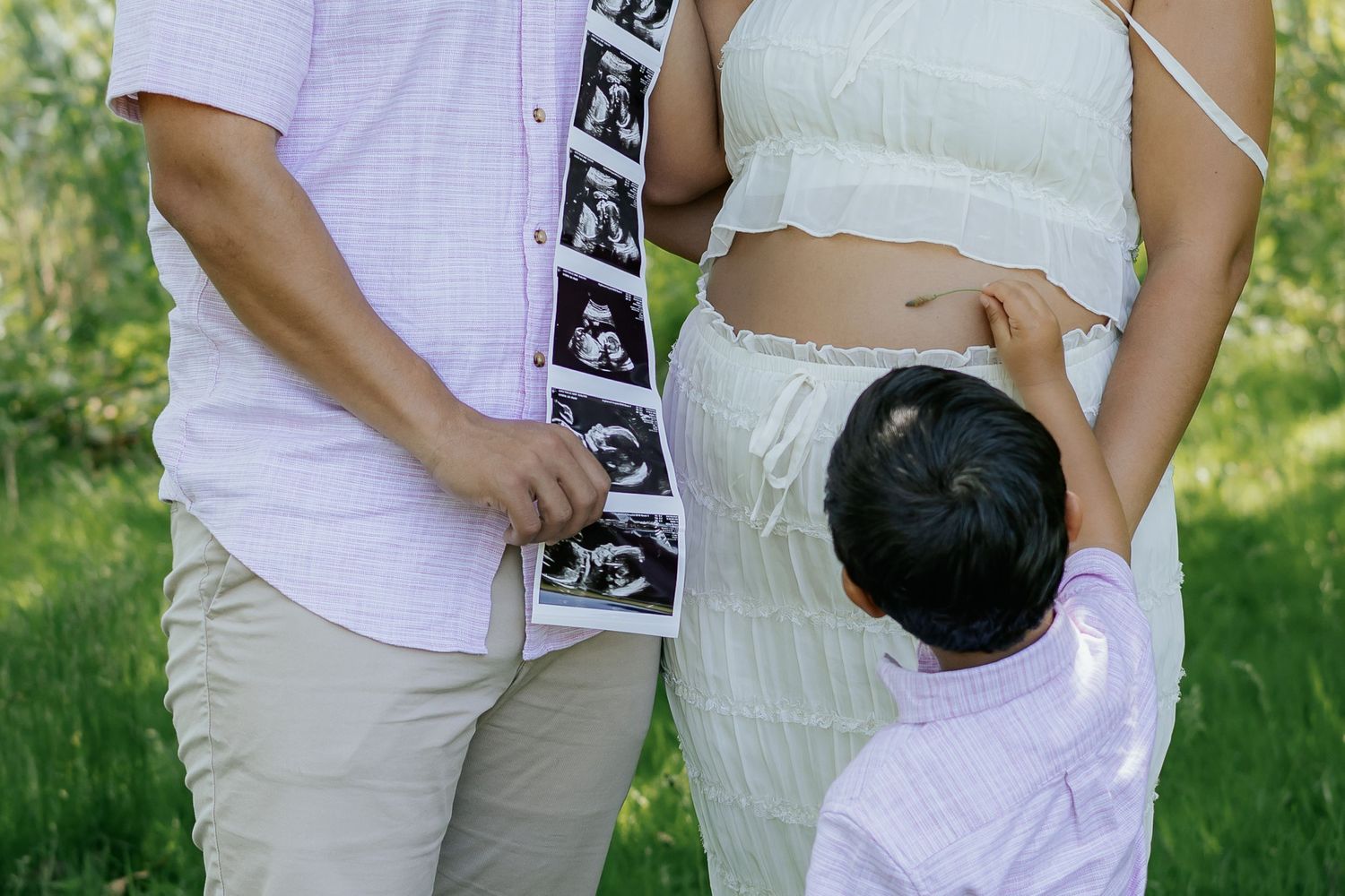 Family maternity photo with parents and young child touching pregnant belly outdoors in green park setting.