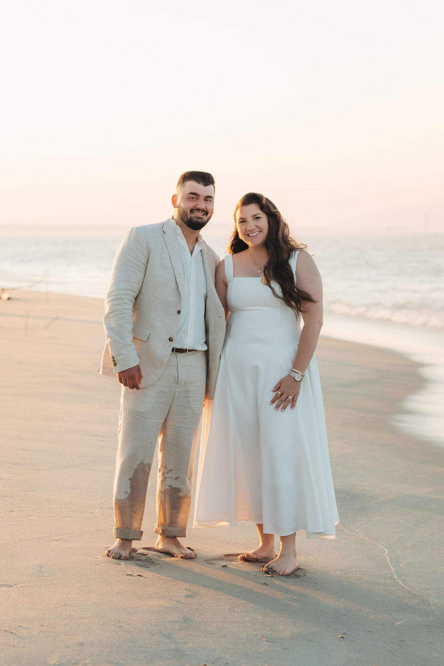 Couple in white dress and beige suit posing barefoot on sandy beach at golden sunset.