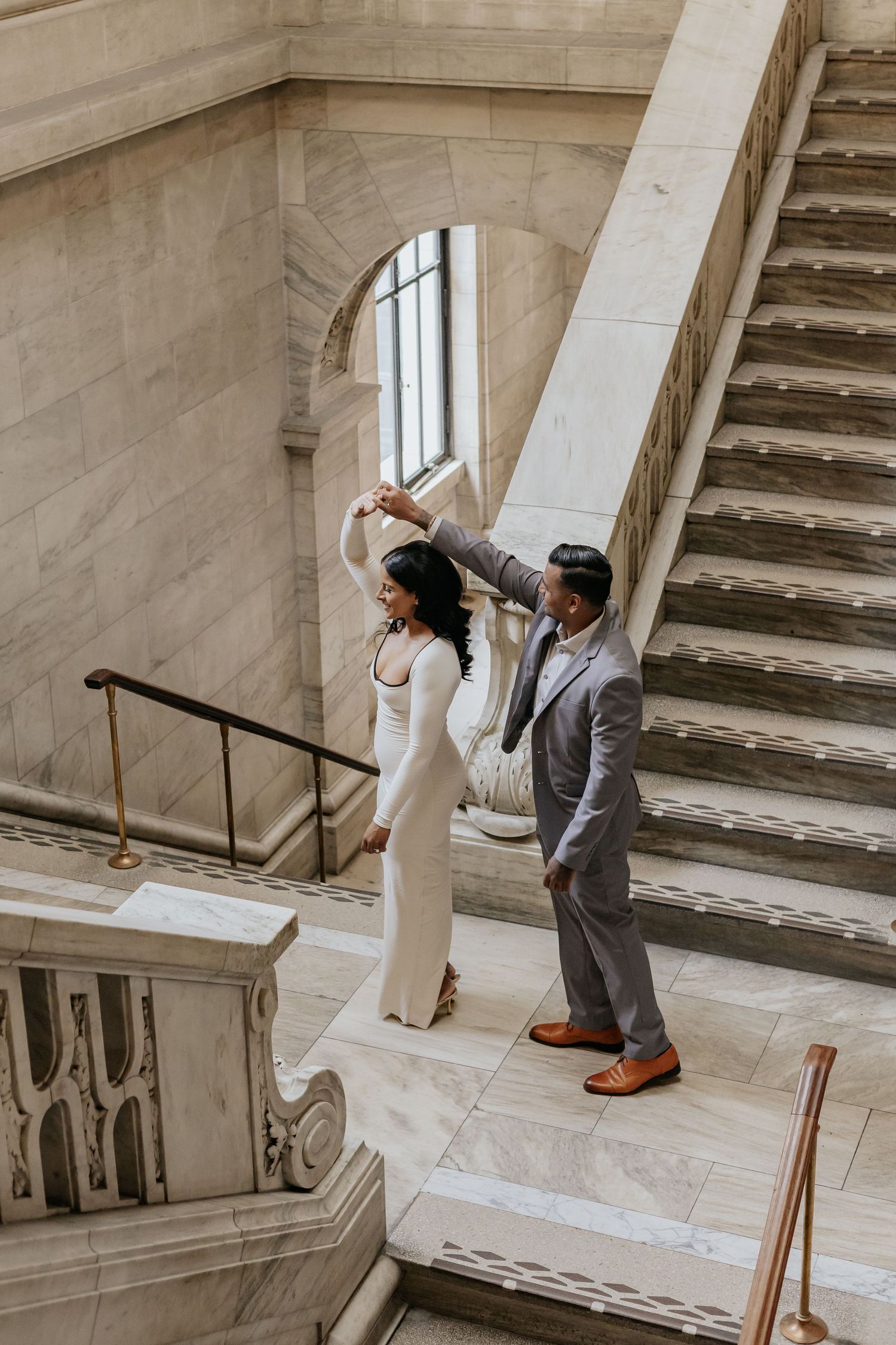 Elegant couple dancing on marble stairs in grand architectural interior, woman in white dress.