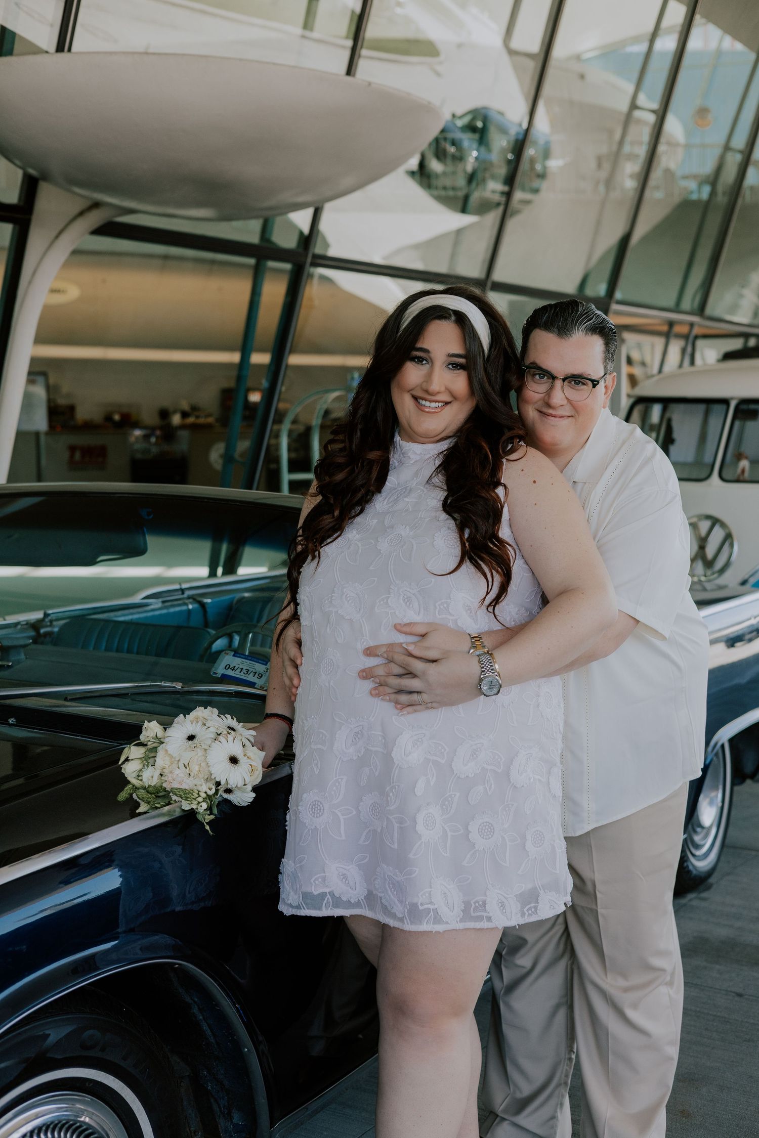 Pregnant bride in white lace dress poses with groom beside vintage black car at TWA Hotel.