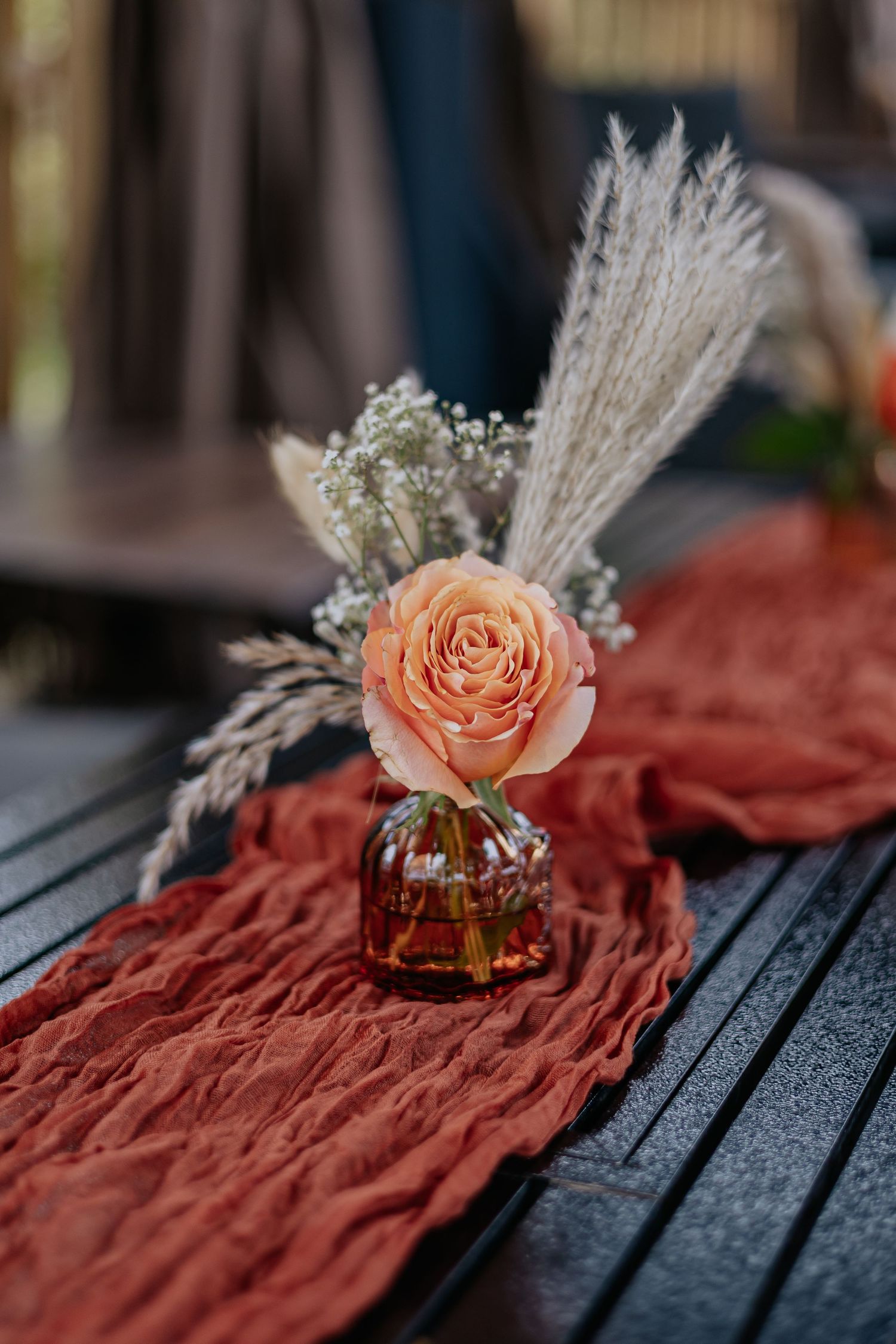 Peach rose with pampas grass in amber vase on rust-colored table runner.