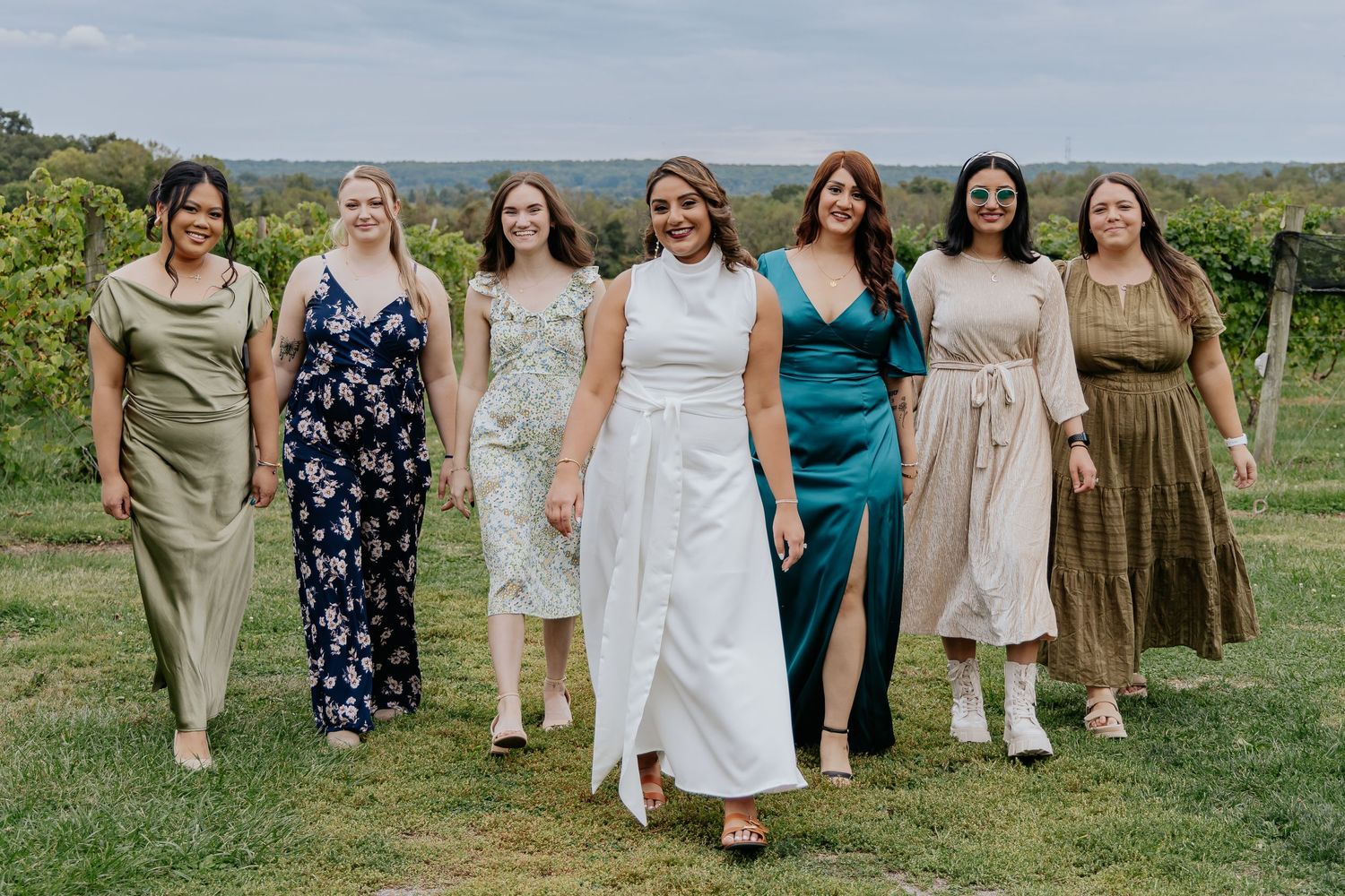 Seven elegantly dressed women walking together through a lush green vineyard on a wedding day.