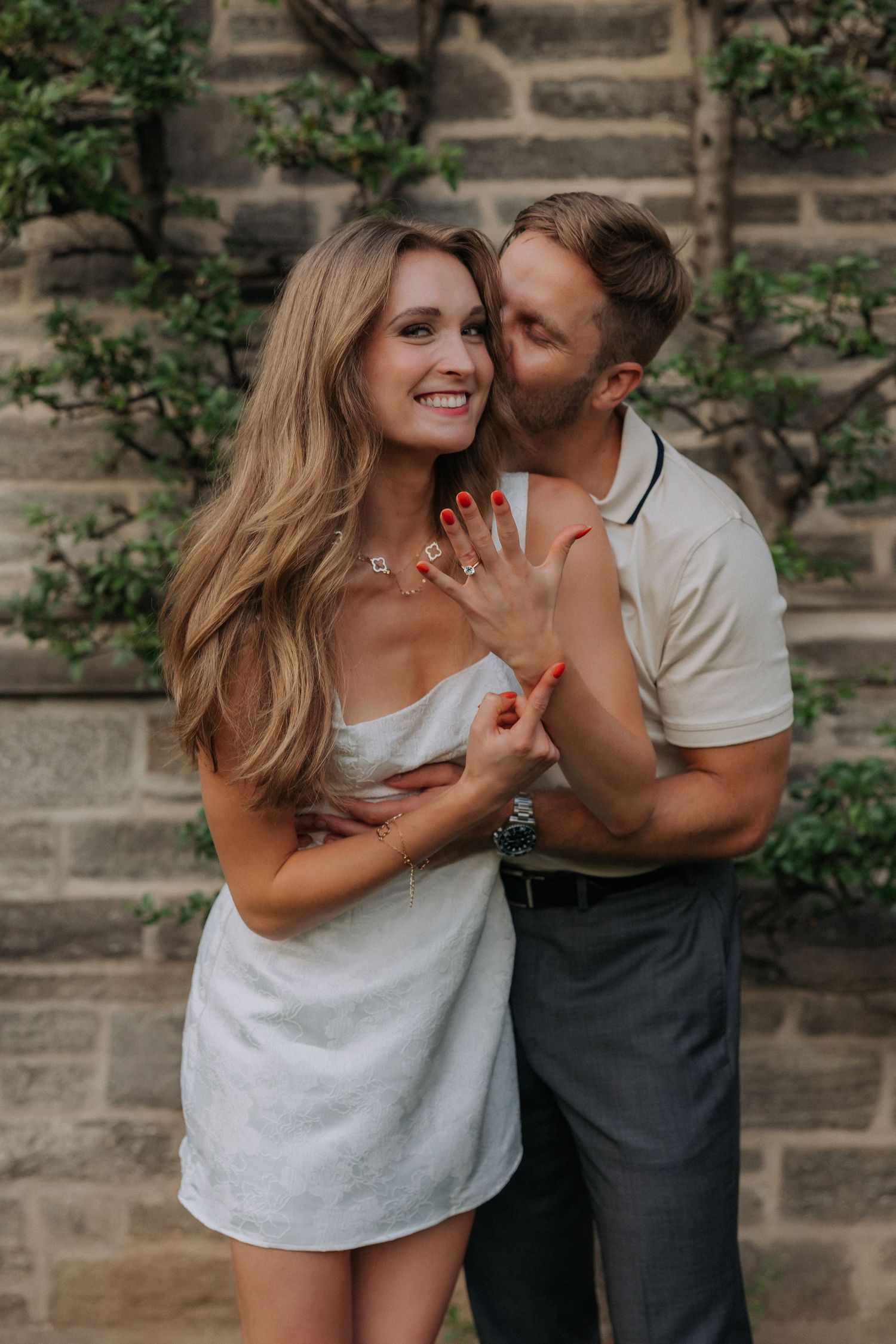Couple embracing outdoors, woman in white mini dress showing engagement ring, man kissing her cheek near stone wall.