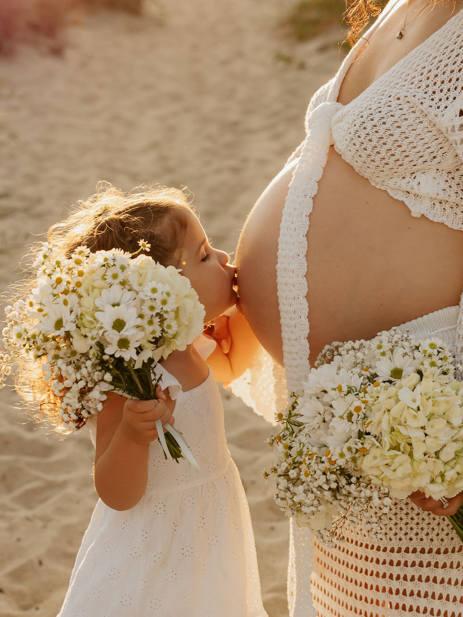 A young girl in white dress kisses pregnant mother's belly, both holding daisy bouquets on a sunny beach.