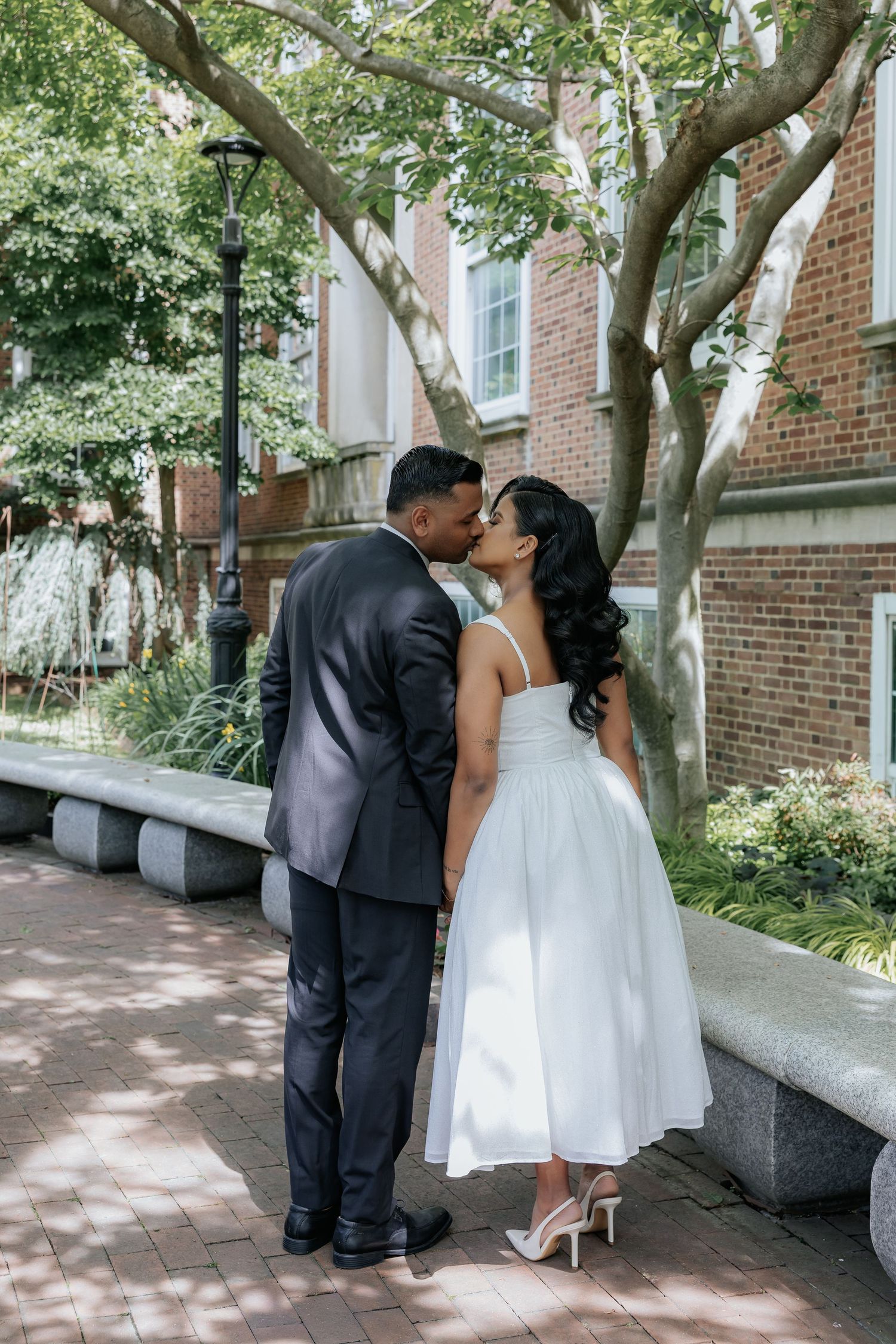 Bride and groom sharing a tender kiss on brick courtyard pathway surrounded by greenery.