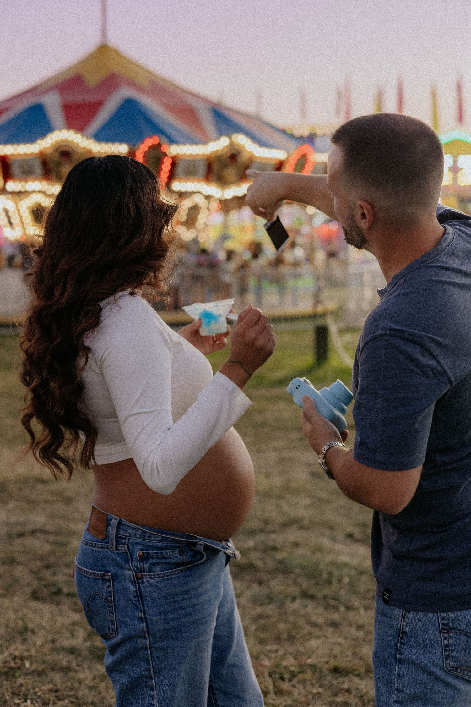 Pregnant couple reviewing photos near vibrant carousel at sunset fair.